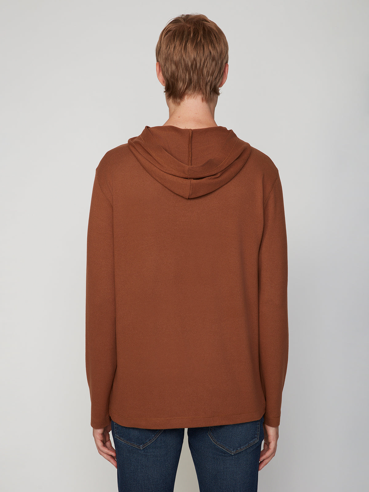A person with short light brown hair wears the PROJEK RAW MENS HOODED LONG SLEEVE KNIT in brown and dark blue jeans, standing with their back to the camera against a light background, evoking a relaxed, casual style.