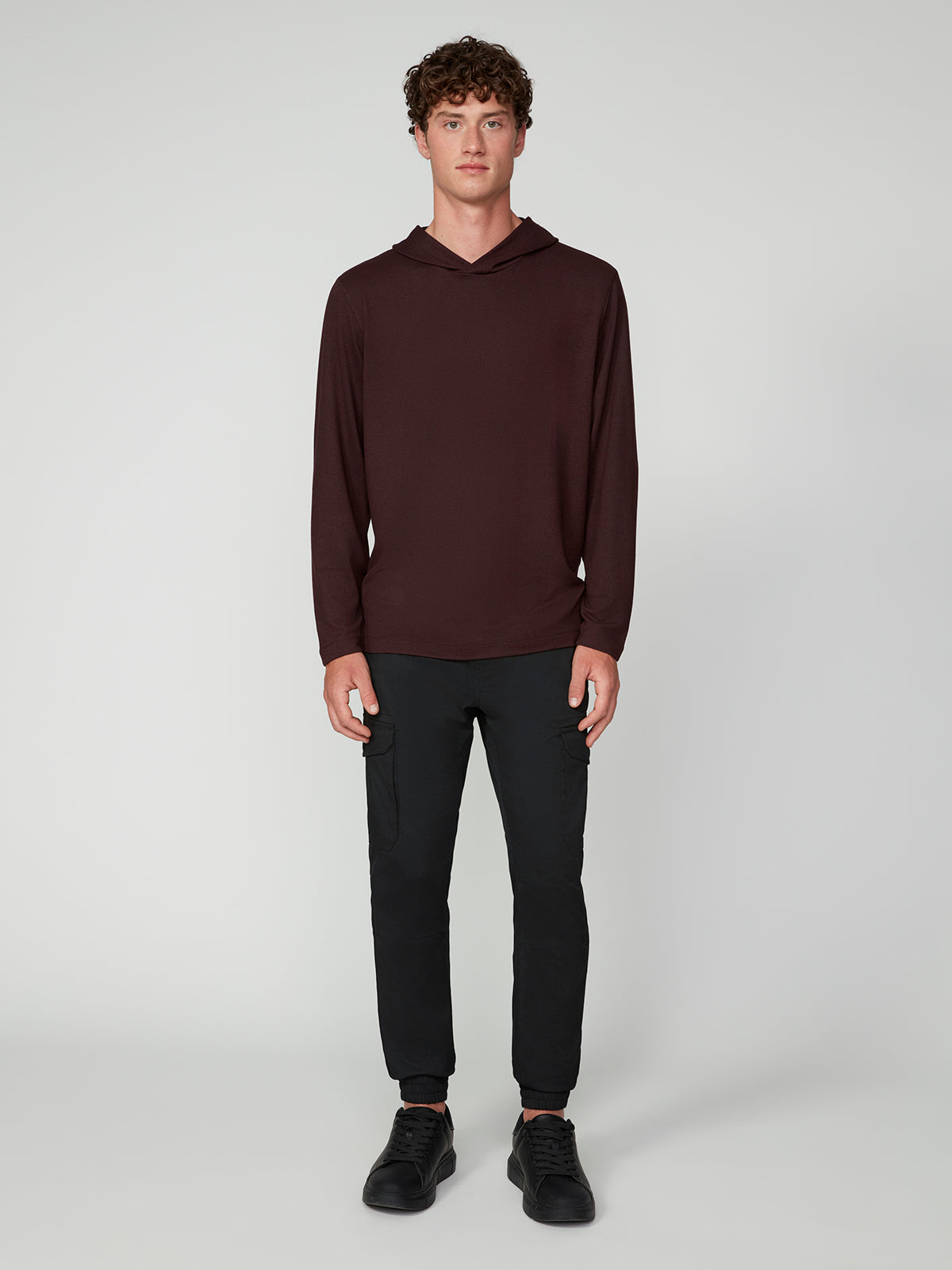 A young man with curly hair faces forward, wearing a maroon PROJEK RAW MENS HOODED LONG SLEEVE KNIT, black pants, and black shoes against a plain light gray background.