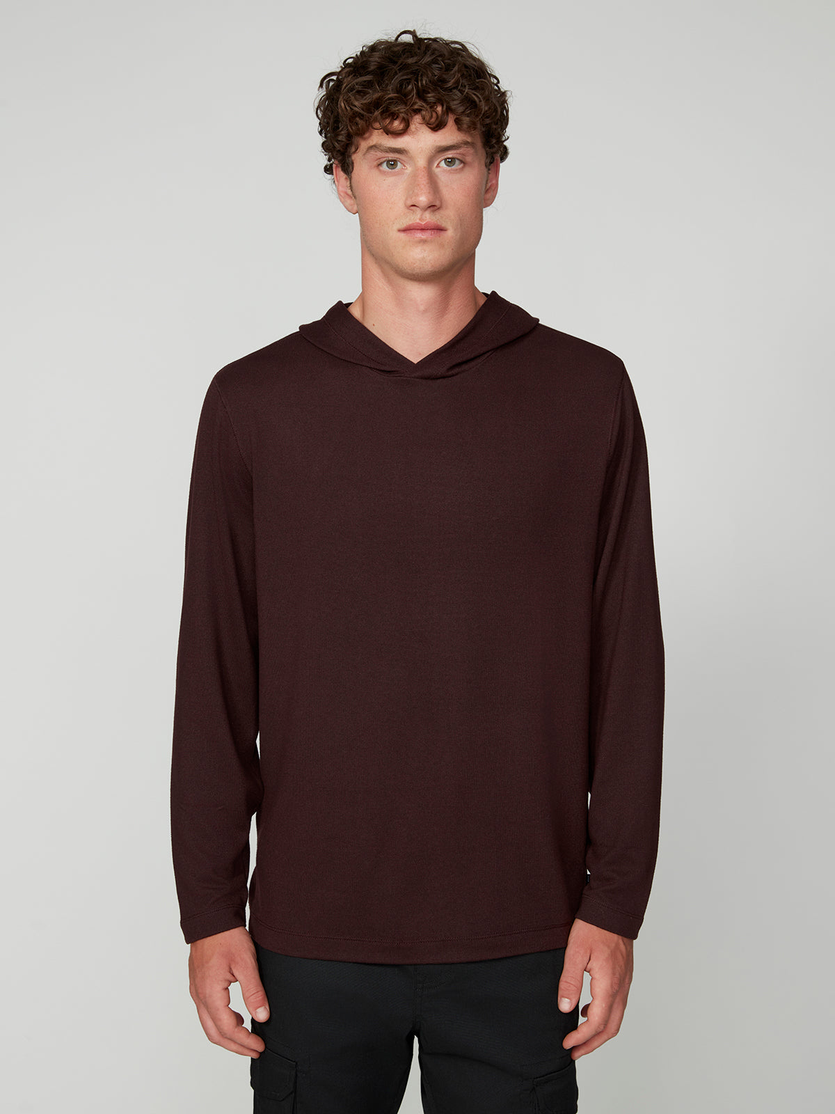 A young man with curly brown hair wears the PROJEK RAW MENS HOODED LONG SLEEVE KNIT in dark maroon and black pants, standing against a light gray background and facing forward with a neutral expression.