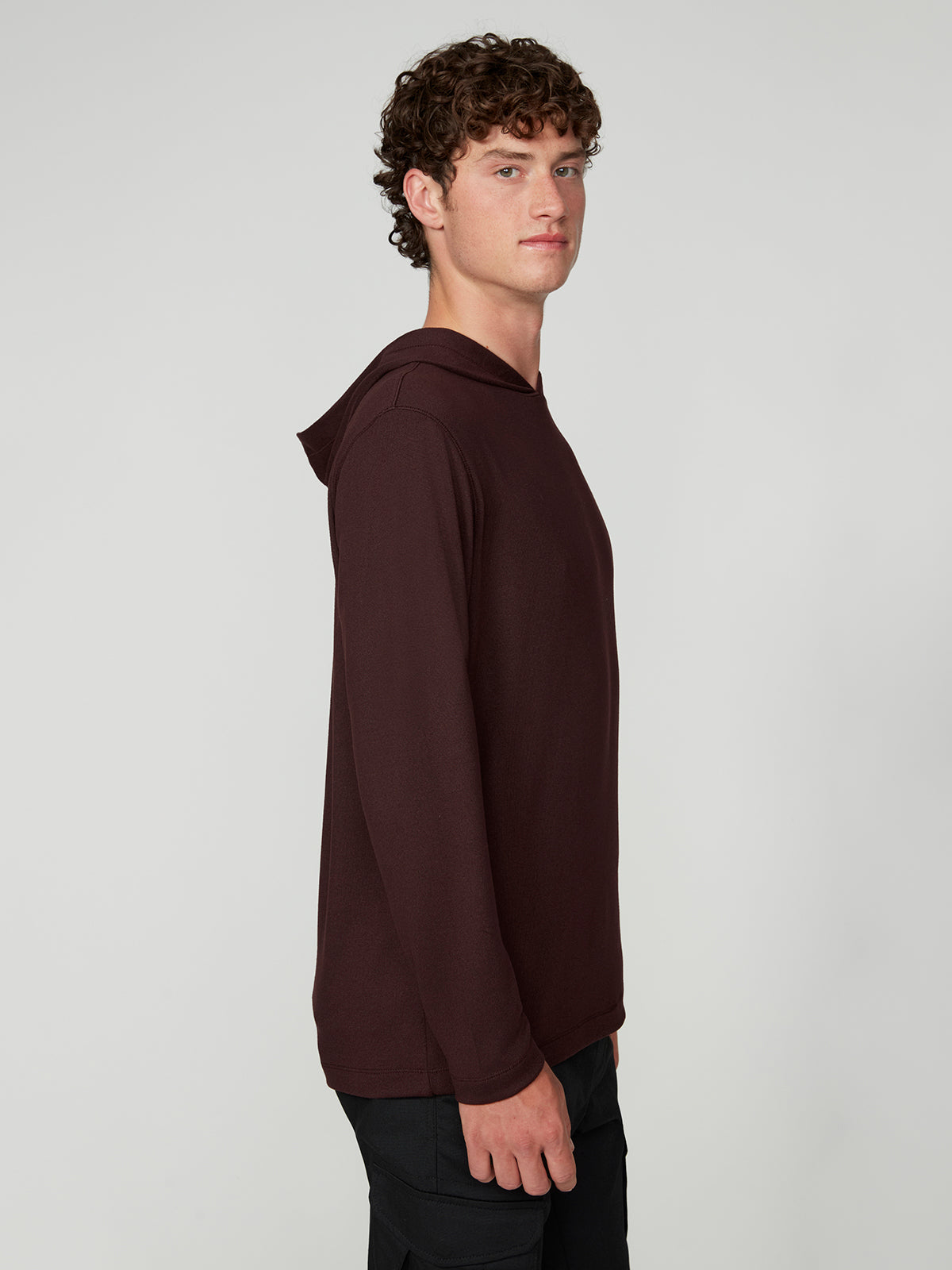 A young man with curly brown hair stands sideways, facing left, wearing a PROJEK RAW MENS HOODED LONG SLEEVE KNIT in dark maroon and black pants against a plain light gray background.