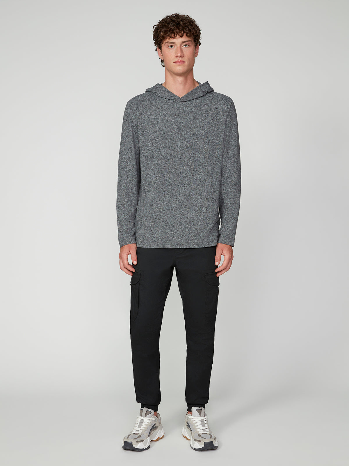 A young man with curly hair faces forward, wearing the PROJEK RAW MENS HOODED LONG SLEEVE KNIT in gray, paired with black jogger pants and white sneakers, embodying casual comfort against a plain light gray background.