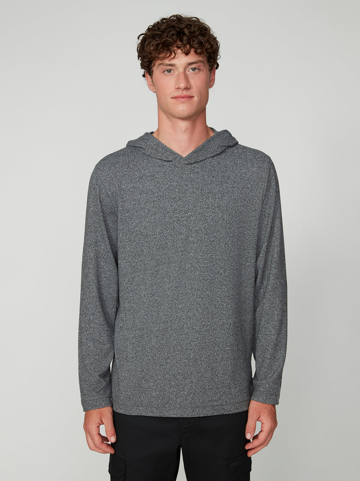 A young man with curly brown hair stands against a plain light gray background, wearing the PROJEK RAW MENS HOODED LONG SLEEVE KNIT. He has a neutral expression and relaxed posture, exuding casual comfort.