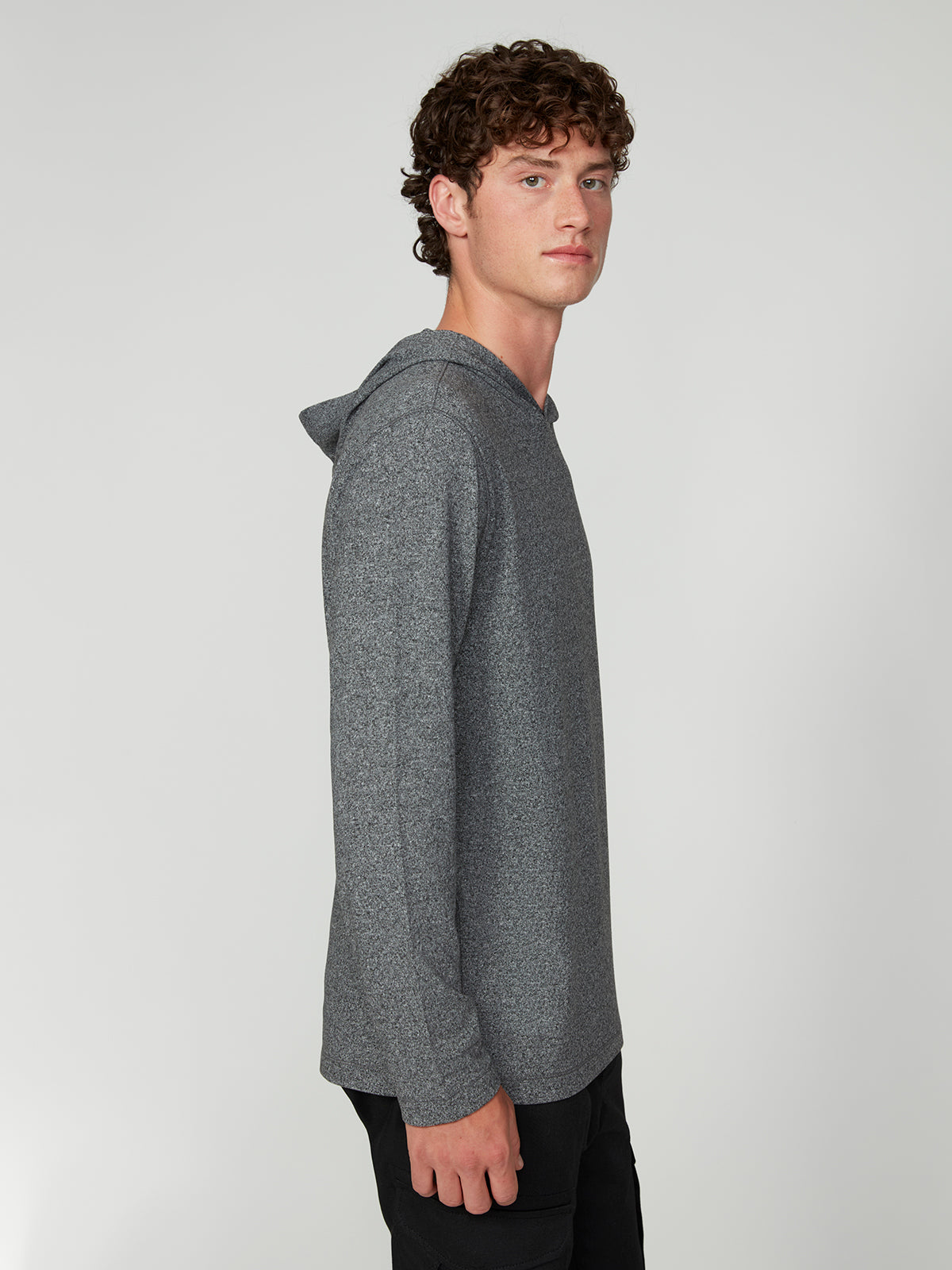 A young man with curly brown hair stands sideways, facing left, wearing the PROJEK RAW MENS HOODED LONG SLEEVE KNIT in gray and black pants, showcasing casual comfort against a plain light gray background.