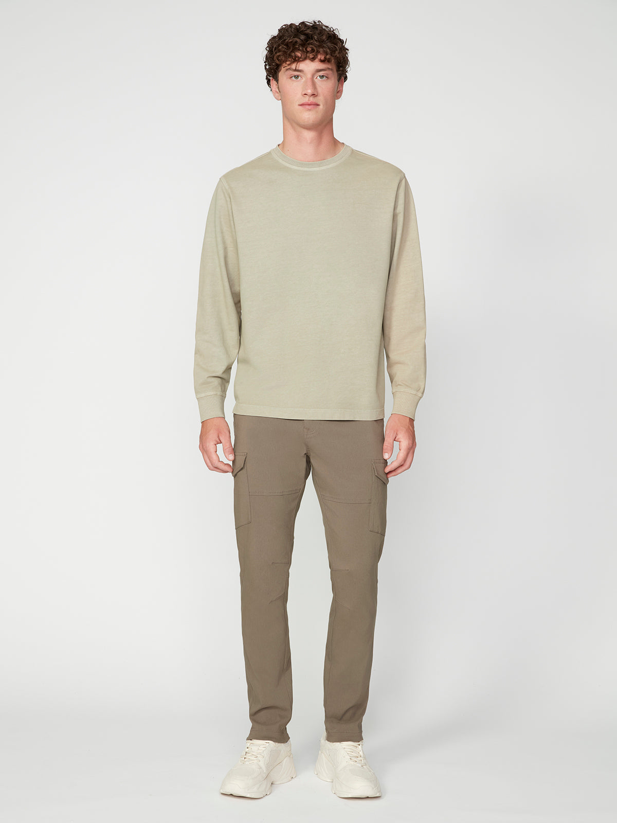 A young man with curly hair stands facing forward, wearing a PROJEK RAW MENS LONG SLEEVE CREW NECK KNIT in light beige French terry, paired with taupe cargo pants and white sneakers against a plain white background.