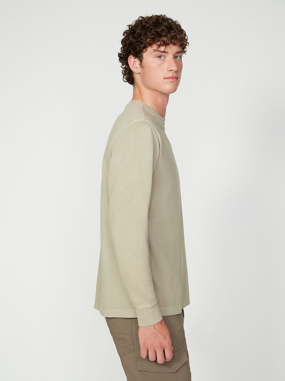 A young man with curly hair, dressed in a PROJEK RAW MENS LONG SLEEVE CREW NECK KNIT and khaki pants, stands in profile against a plain white backdrop, showcasing a relaxed and modern style.