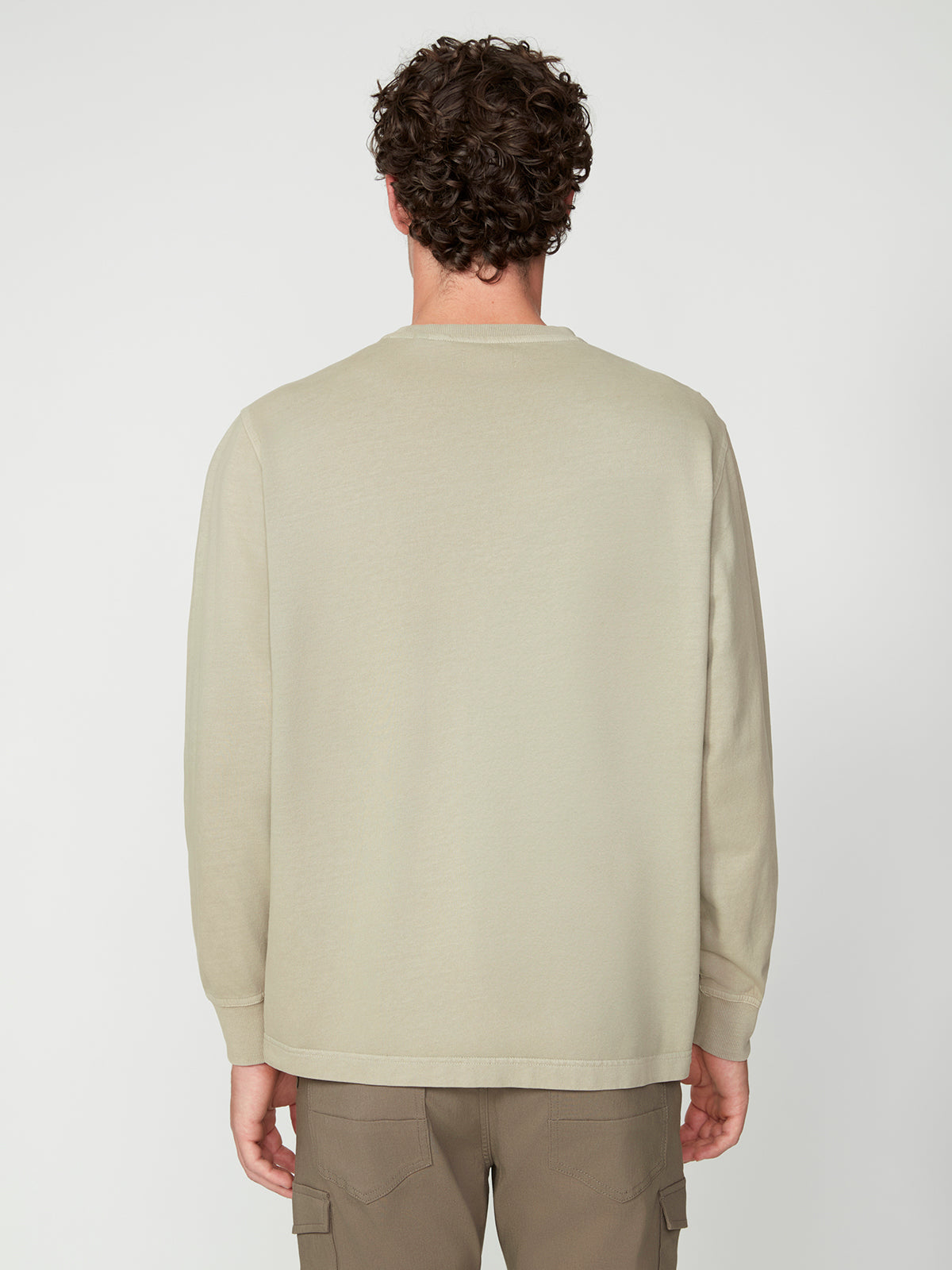 A man with curly brown hair is seen from behind, wearing a PROJEK RAW MENS LONG SLEEVE CREW NECK KNIT in light beige, paired with khaki cargo pants, set against a plain white background.