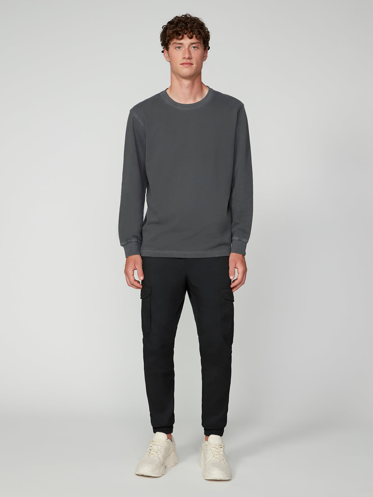 A young man with curly hair faces forward in a casual look, wearing a PROJEK RAW MENS LONG SLEEVE CREW NECK KNIT, black jogger pants, and white sneakers against a plain light gray background.