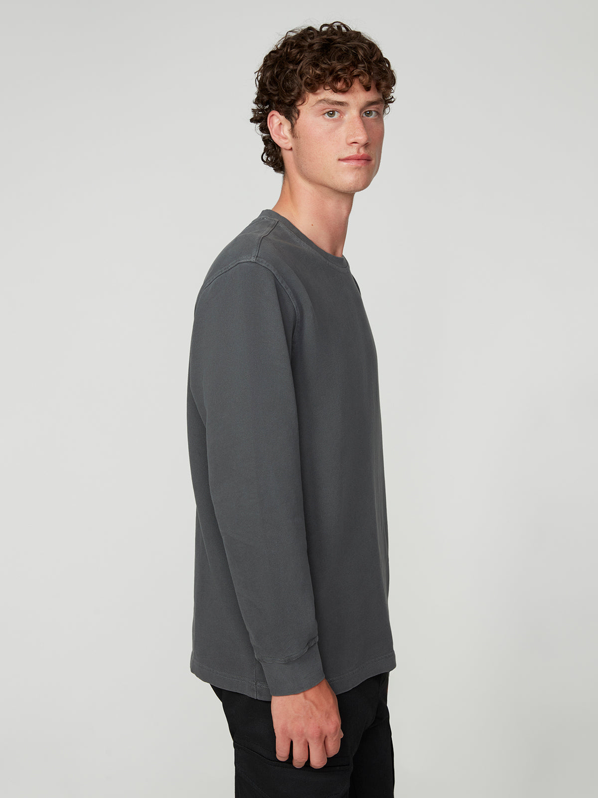 A young man with curly brown hair stands sideways, facing left. He wears the PROJEK RAW MENS LONG SLEEVE CREW NECK KNIT in charcoal gray and black pants, creating a relaxed, casual look against a light gray background.