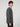 A young man with curly brown hair stands sideways, facing left. He wears the PROJEK RAW MENS LONG SLEEVE CREW NECK KNIT in charcoal gray and black pants, creating a relaxed, casual look against a light gray background.