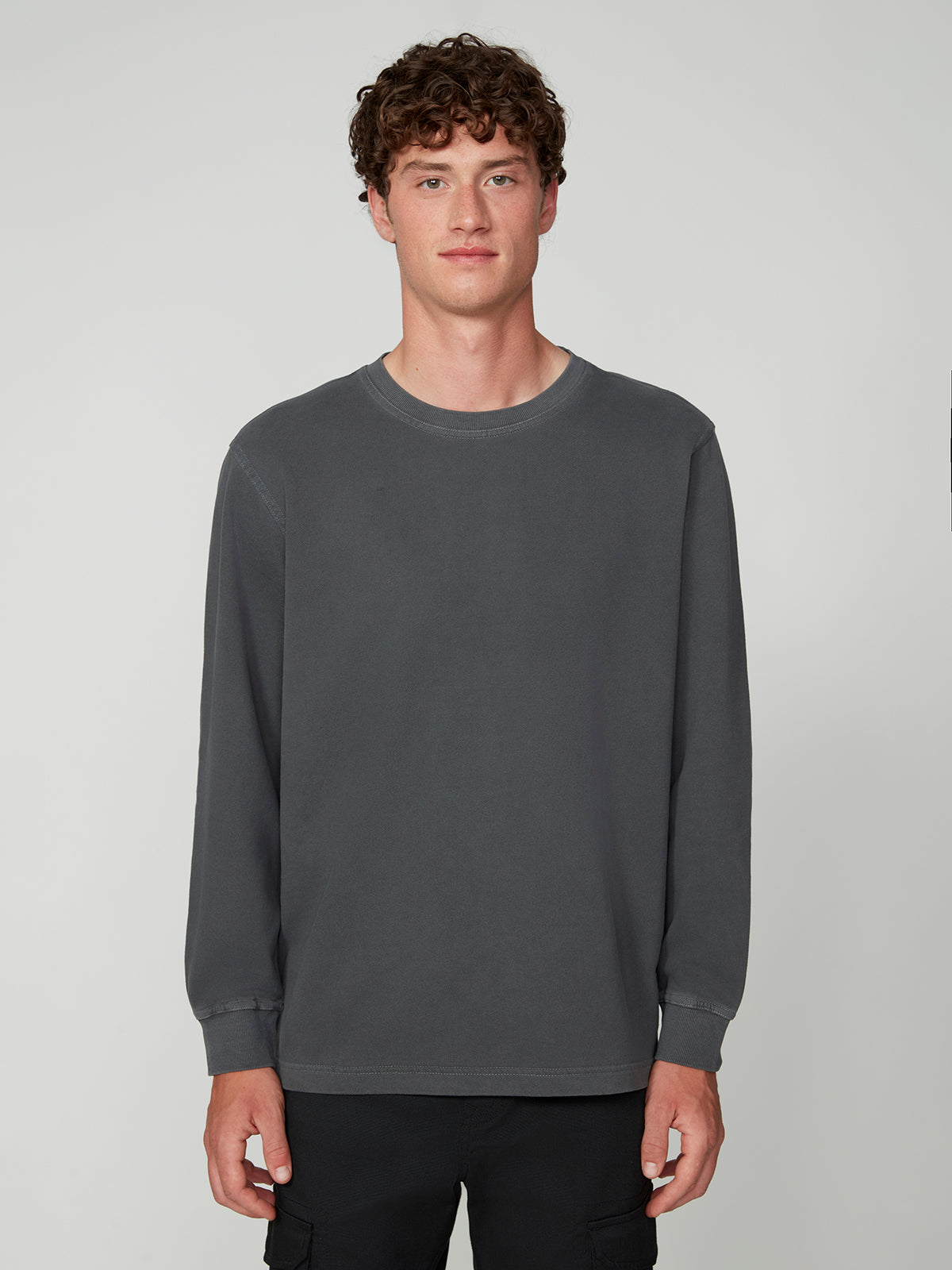A young man with curly brown hair stands against a light gray background, wearing the PROJEK RAW MENS LONG SLEEVE CREW NECK KNIT in dark gray and black pants, looking casually at the camera with a neutral expression.
