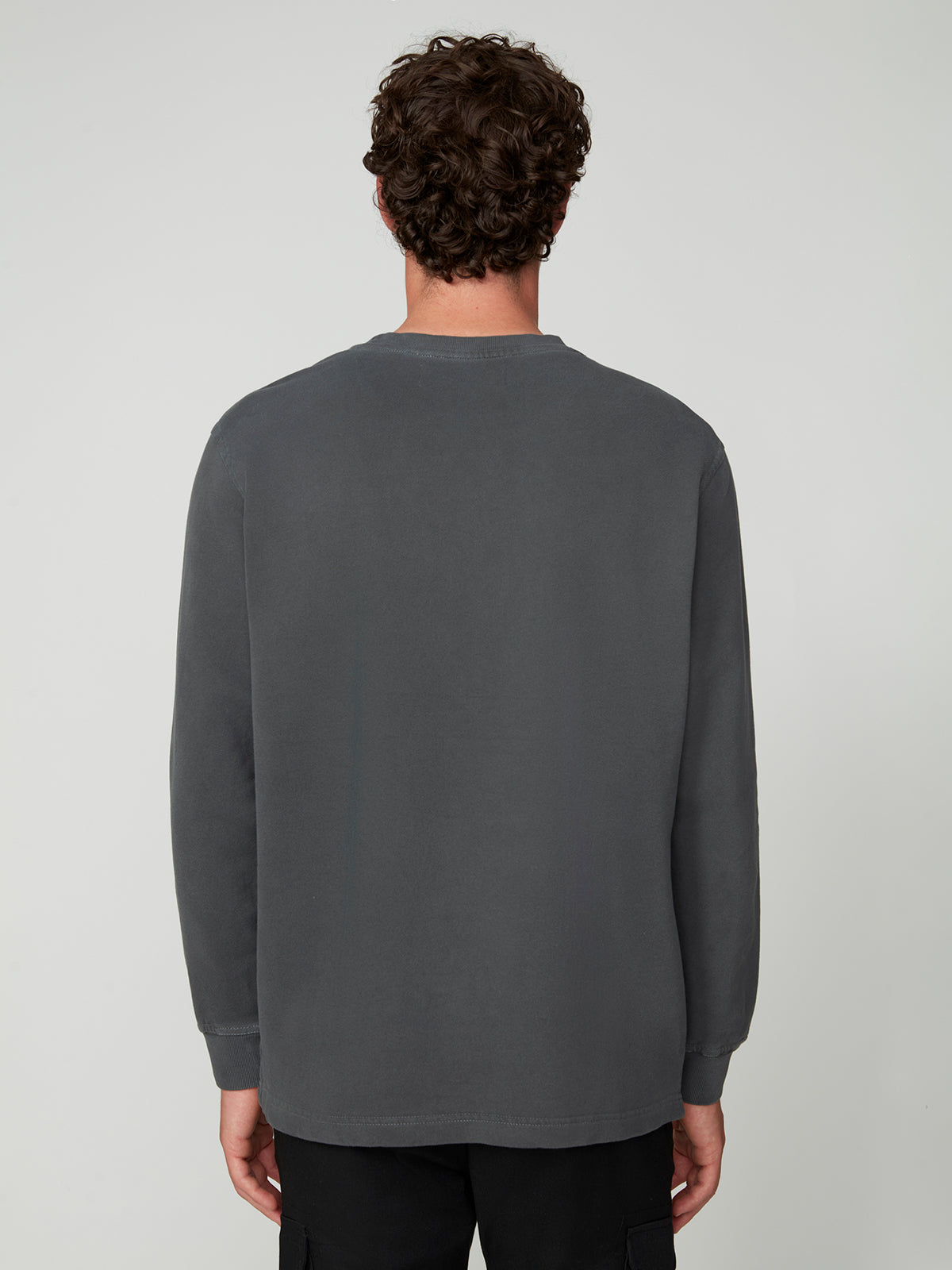 A person with curly brown hair stands with their back to the camera, wearing the PROJEK RAW MENS LONG SLEEVE CREW NECK KNIT and black pants, creating a relaxed, casual look against a plain light gray background.
