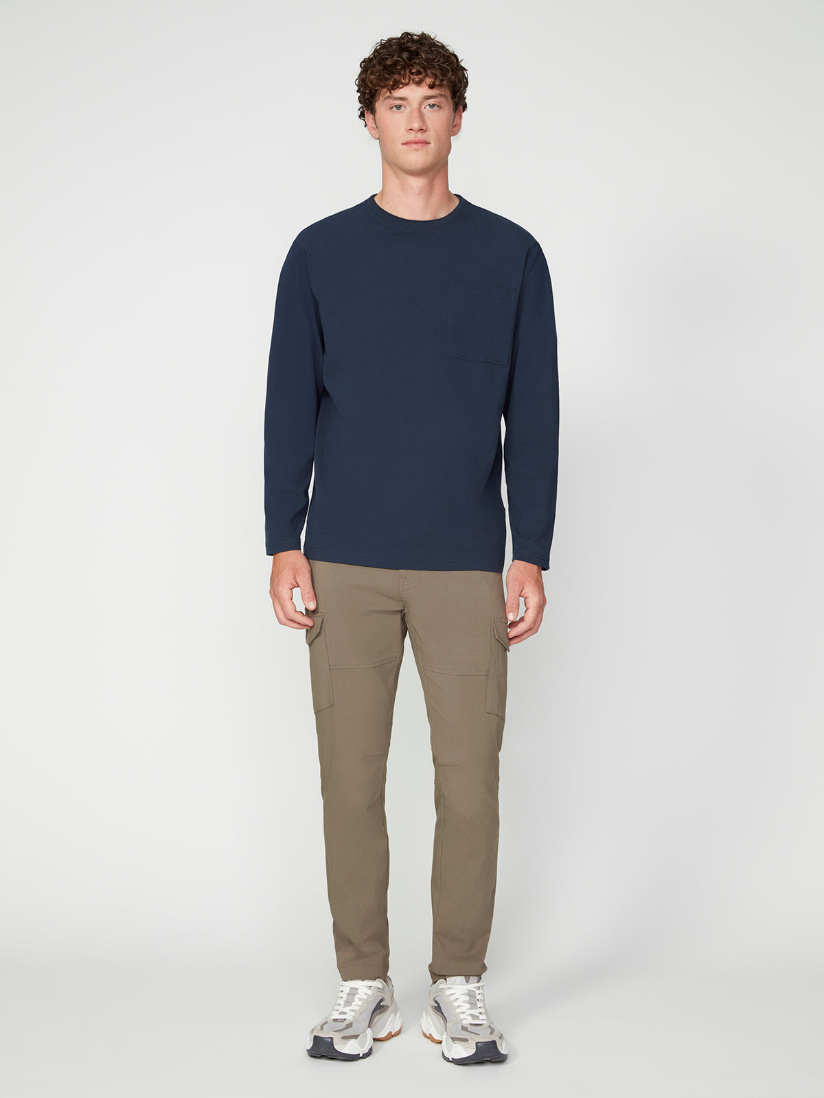 A young man with curly hair stands facing forward, wearing a PROJEK RAW MENS LONG SLEEVE CREW NECK in navy, khaki cargo pants, and white sneakers against a plain light background.