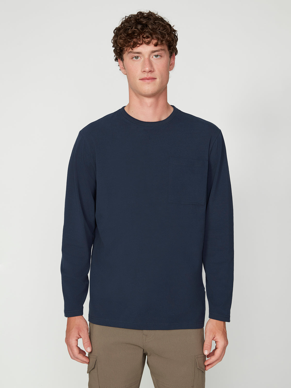 A young man with curly brown hair models the PROJEK RAW MENS LONG SLEEVE CREW NECK, a classic fit navy blue carbon peached jersey shirt with chest pocket, paired with khaki pants against a simple light gray background.