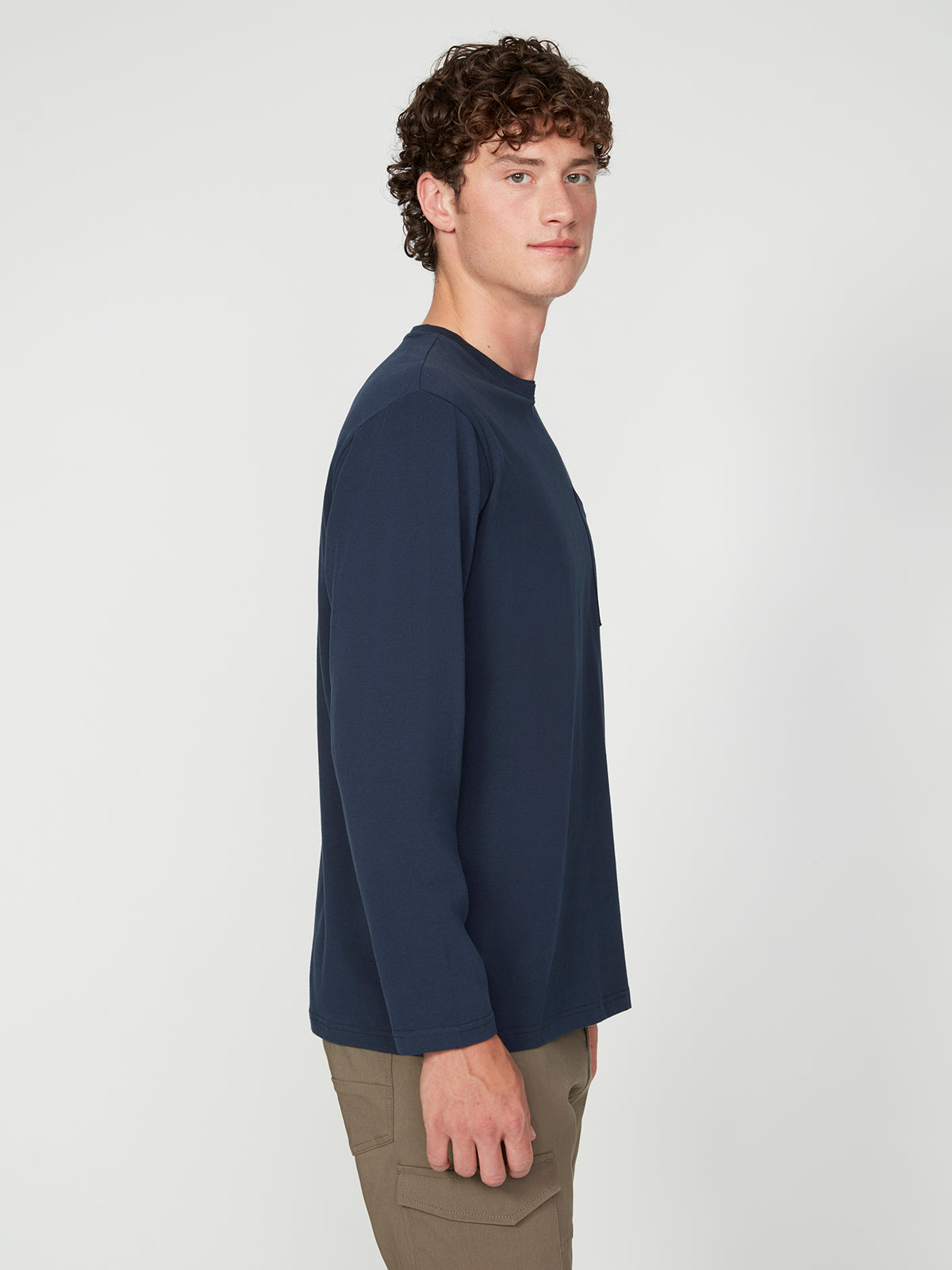 A young man with curly brown hair wears a PROJEK RAW MENS LONG SLEEVE CREW NECK shirt in navy blue and brown pants, standing in profile against a plain white background.
