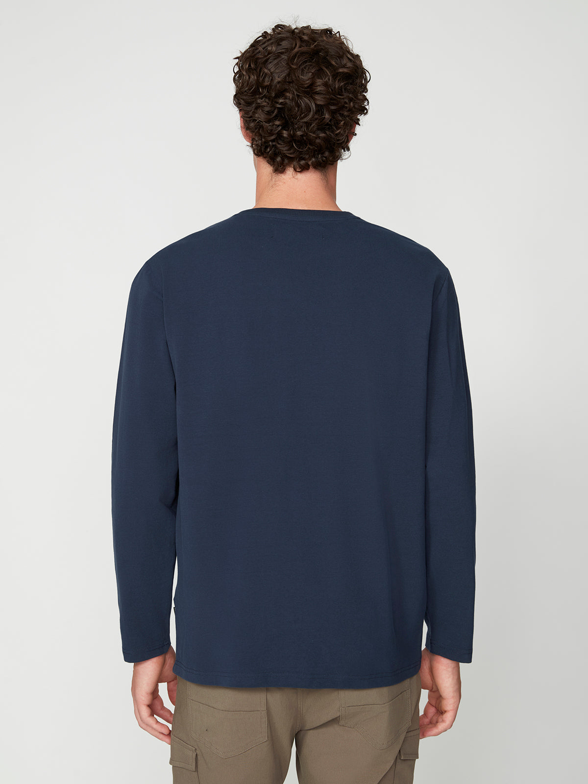 A man with curly brown hair is seen from the back, wearing a PROJEK RAW MENS LONG SLEEVE CREW NECK in navy blue and khaki pants, standing against a plain light background.