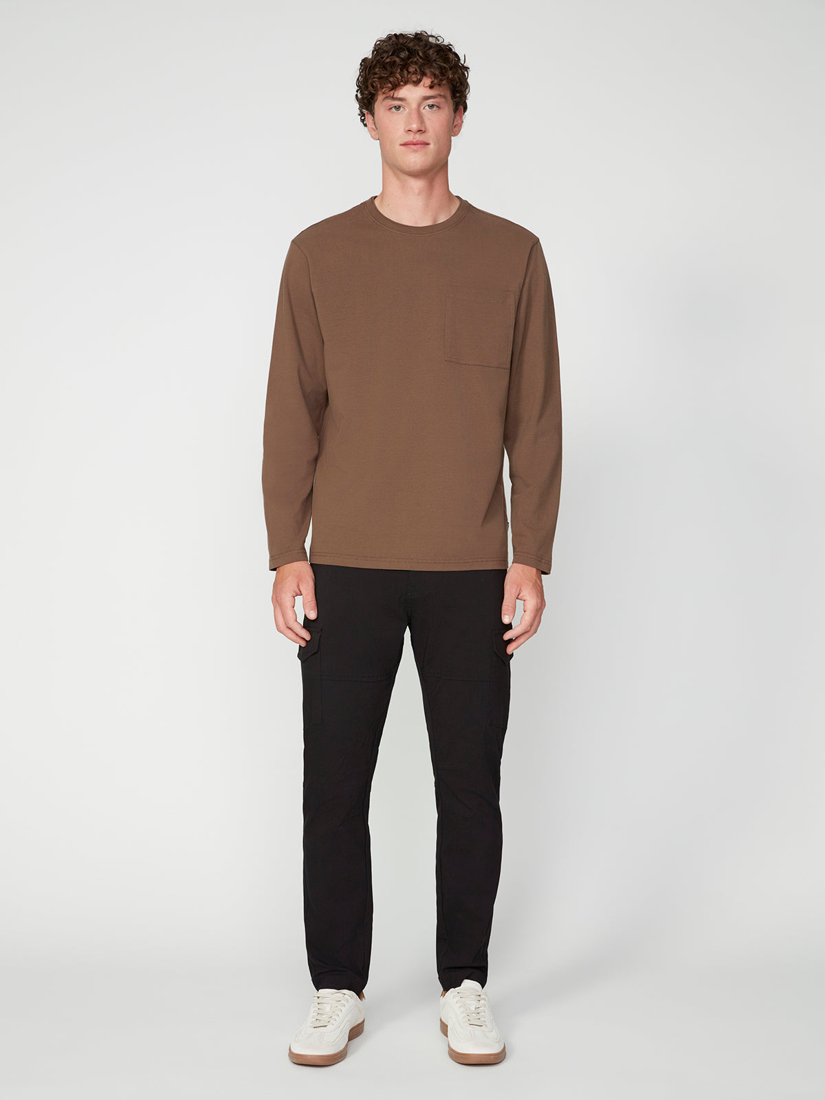 A young man faces forward against a white background, wearing the PROJEK RAW MENS LONG SLEEVE CREW NECK shirt with a chest pocket, paired with black pants and white sneakers.