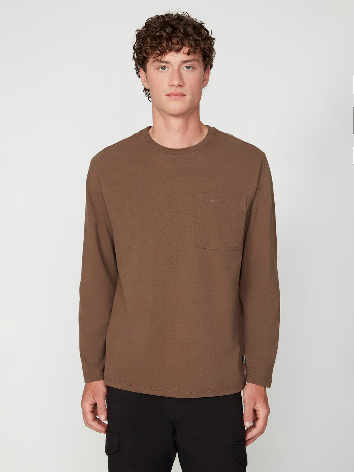 A young man with curly brown hair wears a PROJEK RAW MENS LONG SLEEVE CREW NECK in plain brown, classic fit with a chest pocket and black pants, standing against a light background, facing forward with a neutral expression.