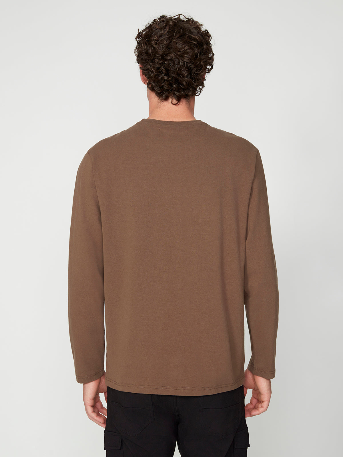 A man with curly brown hair, wearing PROJEK RAW MENS LONG SLEEVE CREW NECK in classic fit and black pants, stands facing away from the camera against a plain light background.