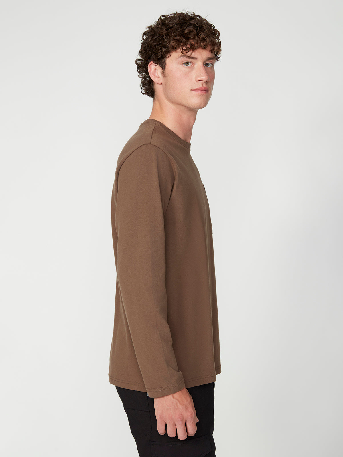 A young man with curly brown hair stands sideways, facing left, wearing a PROJEK RAW MENS LONG SLEEVE CREW NECK in brown and black pants against a plain, light-colored background.