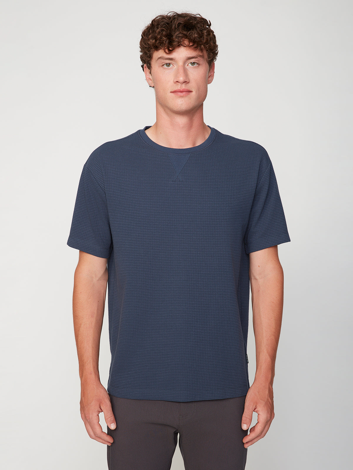 A young man with curly brown hair faces forward, wearing a navy blue PROJEK RAW MENS SHORT SLEEVE CREW NECK GRID JERSEY OVERSIZE TSHIRT and dark pants, set against a plain light background.