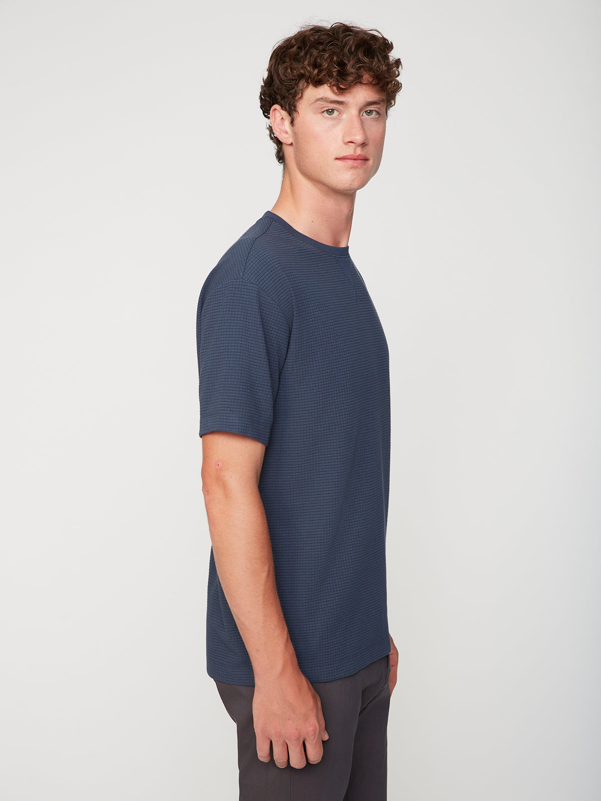 A young man with curly brown hair models the PROJEK RAW MENS SHORT SLEEVE CREW NECK GRID JERSEY OVERSIZE TSHIRT in navy blue and gray pants, standing sideways and looking at the camera against a plain white background.