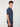 A young man with curly brown hair models the PROJEK RAW MENS SHORT SLEEVE CREW NECK GRID JERSEY OVERSIZE TSHIRT in navy blue and gray pants, standing sideways and looking at the camera against a plain white background.