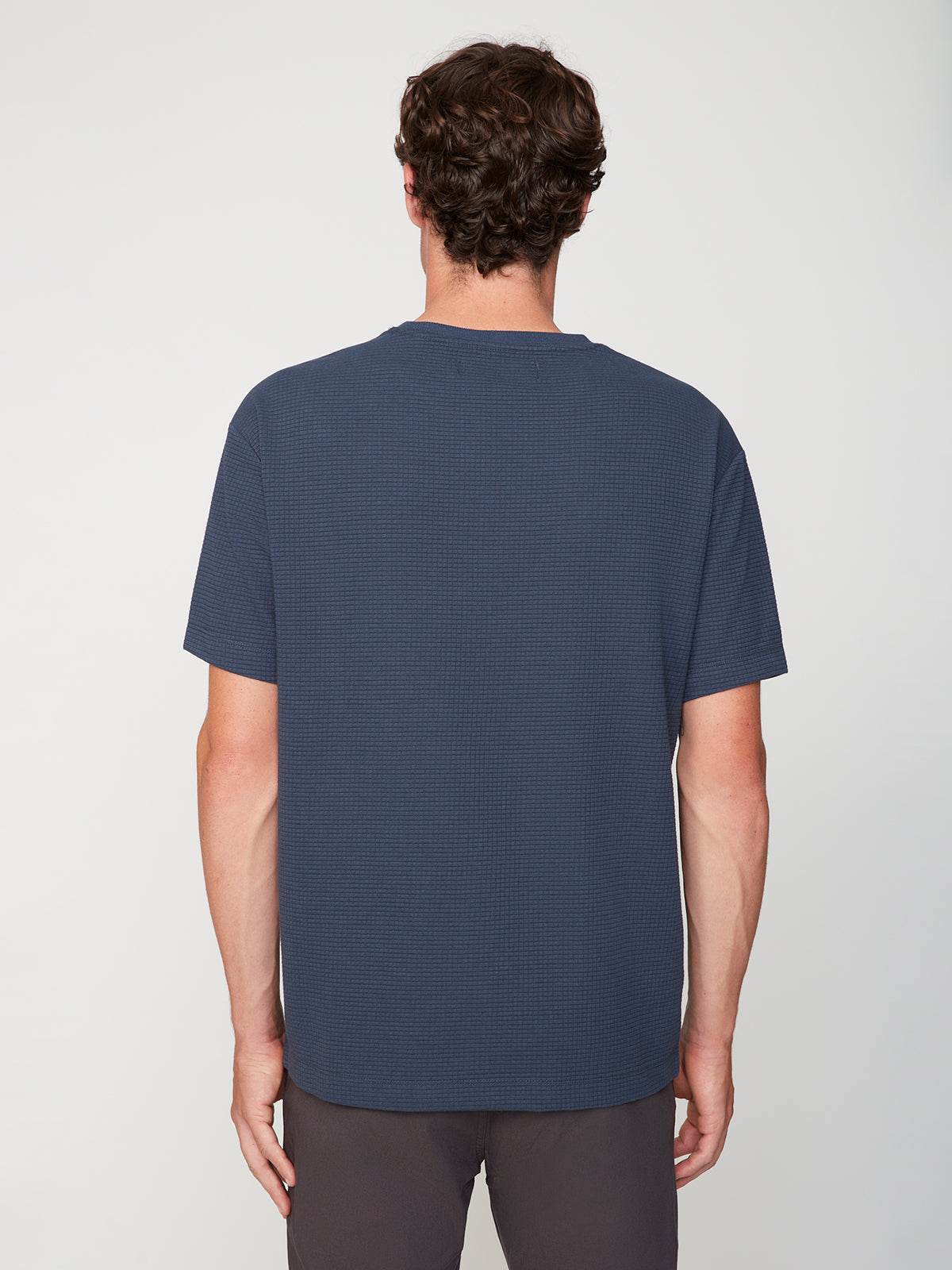 A person with short brown hair stands with their back to the camera, wearing a PROJEK RAW MENS SHORT SLEEVE CREW NECK GRID JERSEY OVERSIZE TSHIRT in dark blue and dark pants against a plain light background.