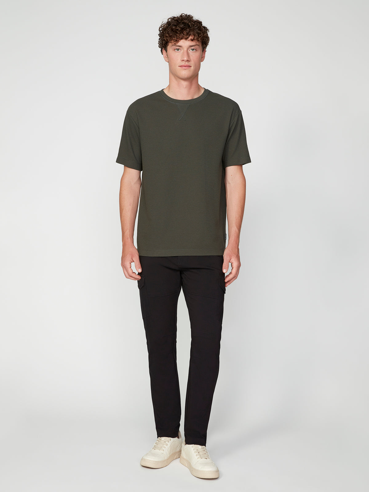 A young man with curly hair faces forward, wearing a PROJEK RAW MENS SHORT SLEEVE CREW NECK GRID JERSEY OVERSIZE TSHIRT, black pants, and white sneakers against a plain light background.