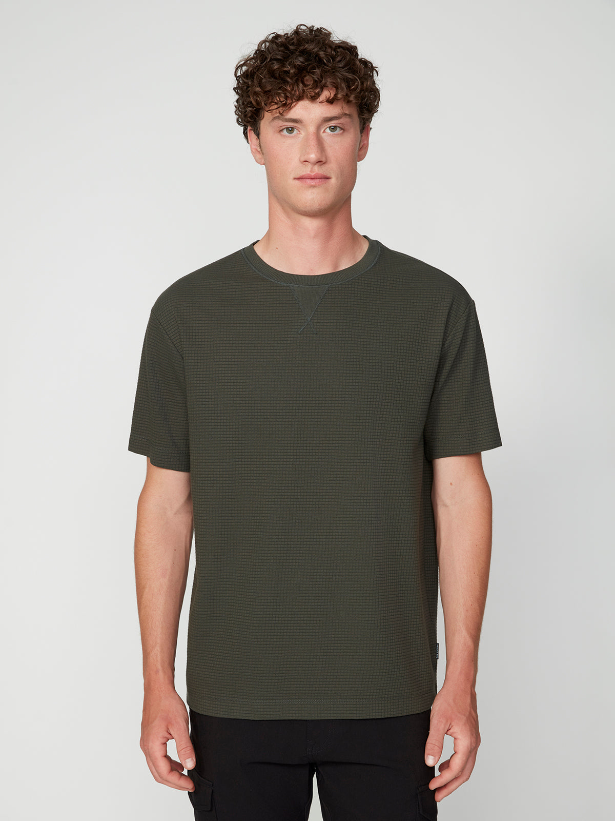 A young man with curly brown hair faces forward against a plain background, wearing a PROJEK RAW MENS SHORT SLEEVE CREW NECK GRID JERSEY OVERSIZE TSHIRT in textured dark green and black pants for a relaxed, casual style.