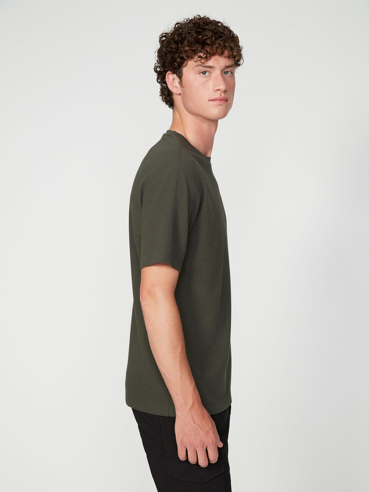 A young man with curly brown hair stands sideways, wearing a PROJEK RAW MENS SHORT SLEEVE CREW NECK GRID JERSEY OVERSIZE TSHIRT and black pants, displaying a relaxed, casual look against a plain light background.