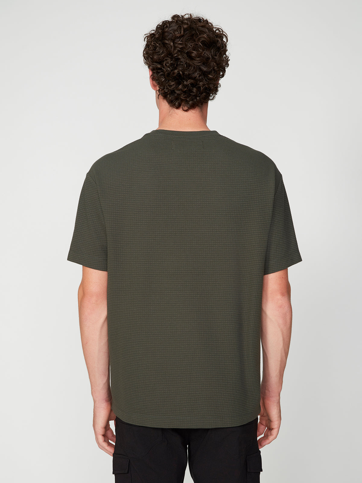 A person with curly hair faces away from the camera, modeling a dark green PROJEK RAW MENS SHORT SLEEVE CREW NECK GRID JERSEY OVERSIZE TSHIRT and black pants against a plain light gray background.