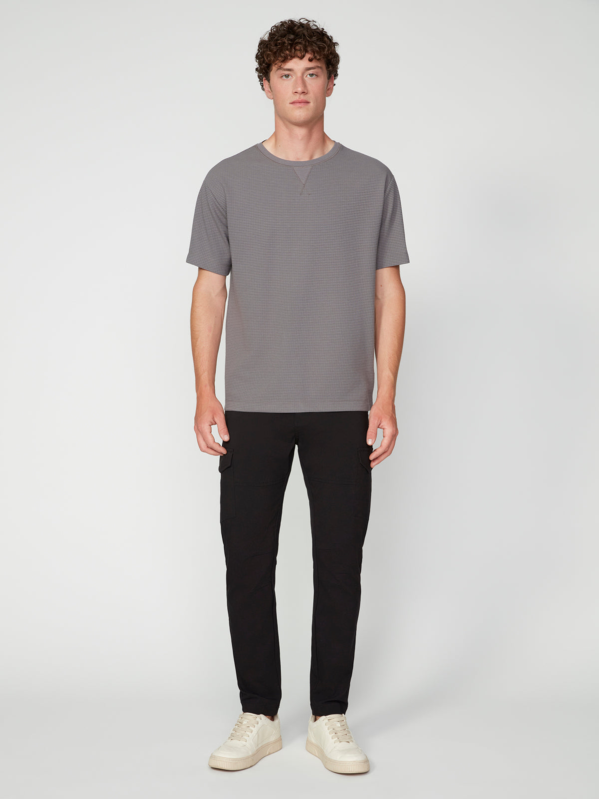 A young man with curly brown hair faces forward in an oversized PROJEK RAW MENS SHORT SLEEVE CREW NECK GRID JERSEY TSHIRT, gray color, paired with black pants and white sneakers, embodying a relaxed casual look against a plain light background.