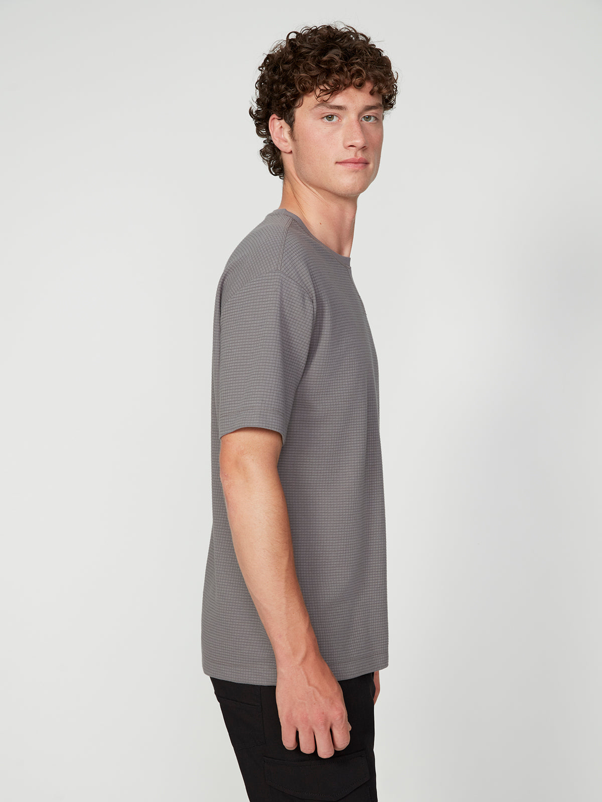 A young man with curly hair stands in profile against a plain background, wearing a PROJEK RAW MENS SHORT SLEEVE CREW NECK GRID JERSEY OVERSIZE TSHIRT in textured gray and black pants, highlighting a relaxed, casual look.