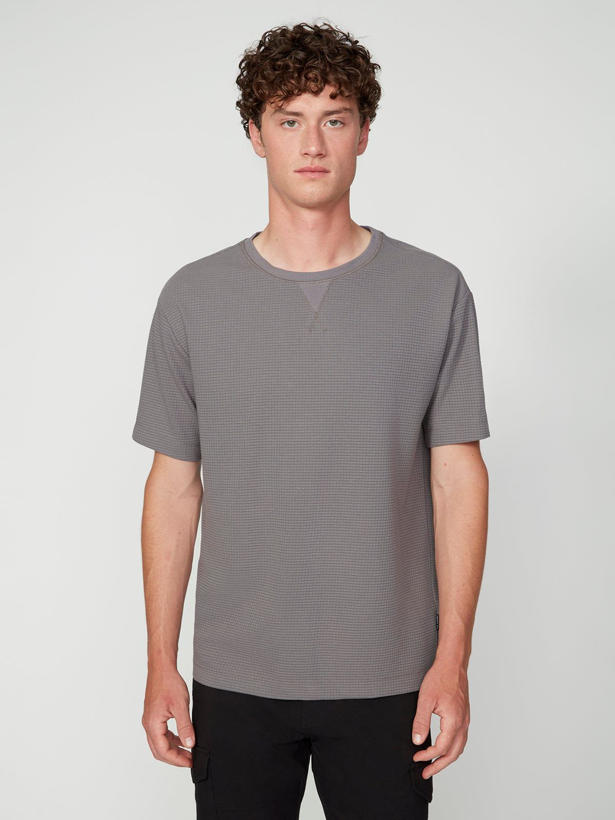 A young man with short curly brown hair wears a PROJEK RAW MENS SHORT SLEEVE CREW NECK GRID JERSEY OVERSIZE TSHIRT and black pants, standing against a light gray background and looking at the camera with a neutral expression.