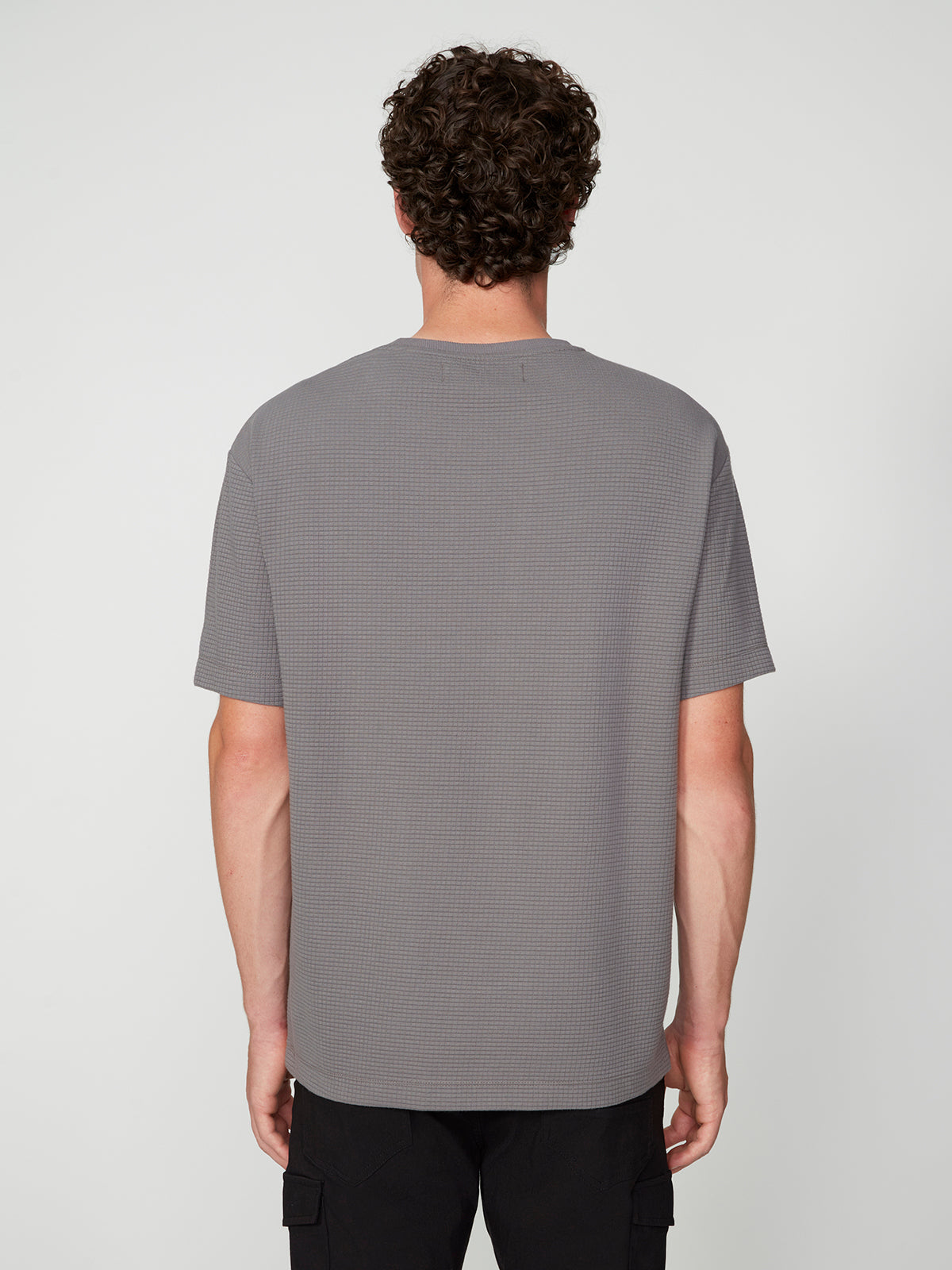 A person with curly brown hair, seen from behind, wears a PROJEK RAW MENS SHORT SLEEVE CREW NECK GRID JERSEY OVERSIZE TSHIRT in textured gray and black pants, showcasing a relaxed style against a plain light background.
