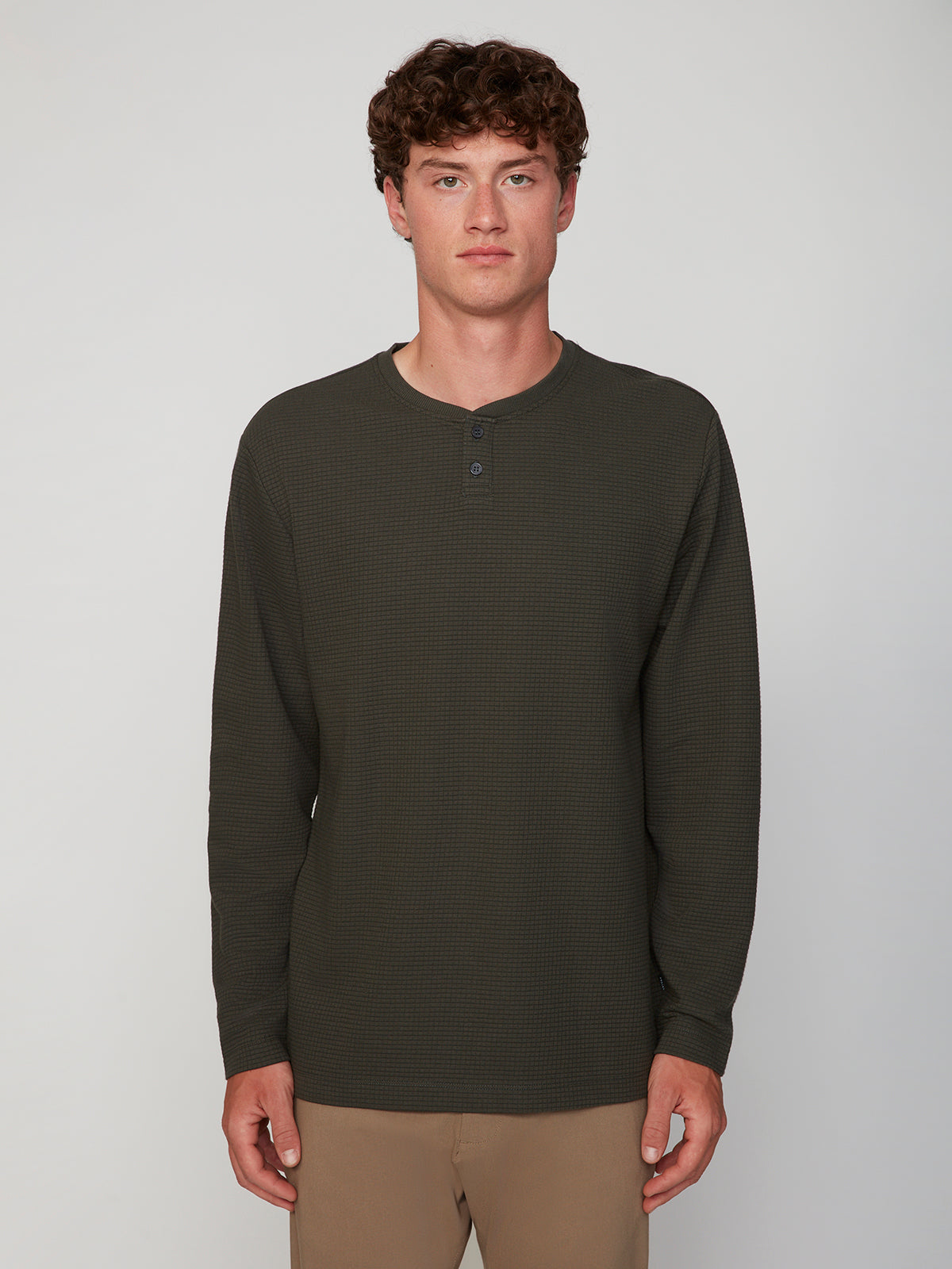 A young man with short curly hair wears a classic fit PROJEK RAW MENS LONG SLEEVE HENLEY GRID JERSEY in dark green and tan pants, standing against a plain light gray background and facing the camera neutrally.