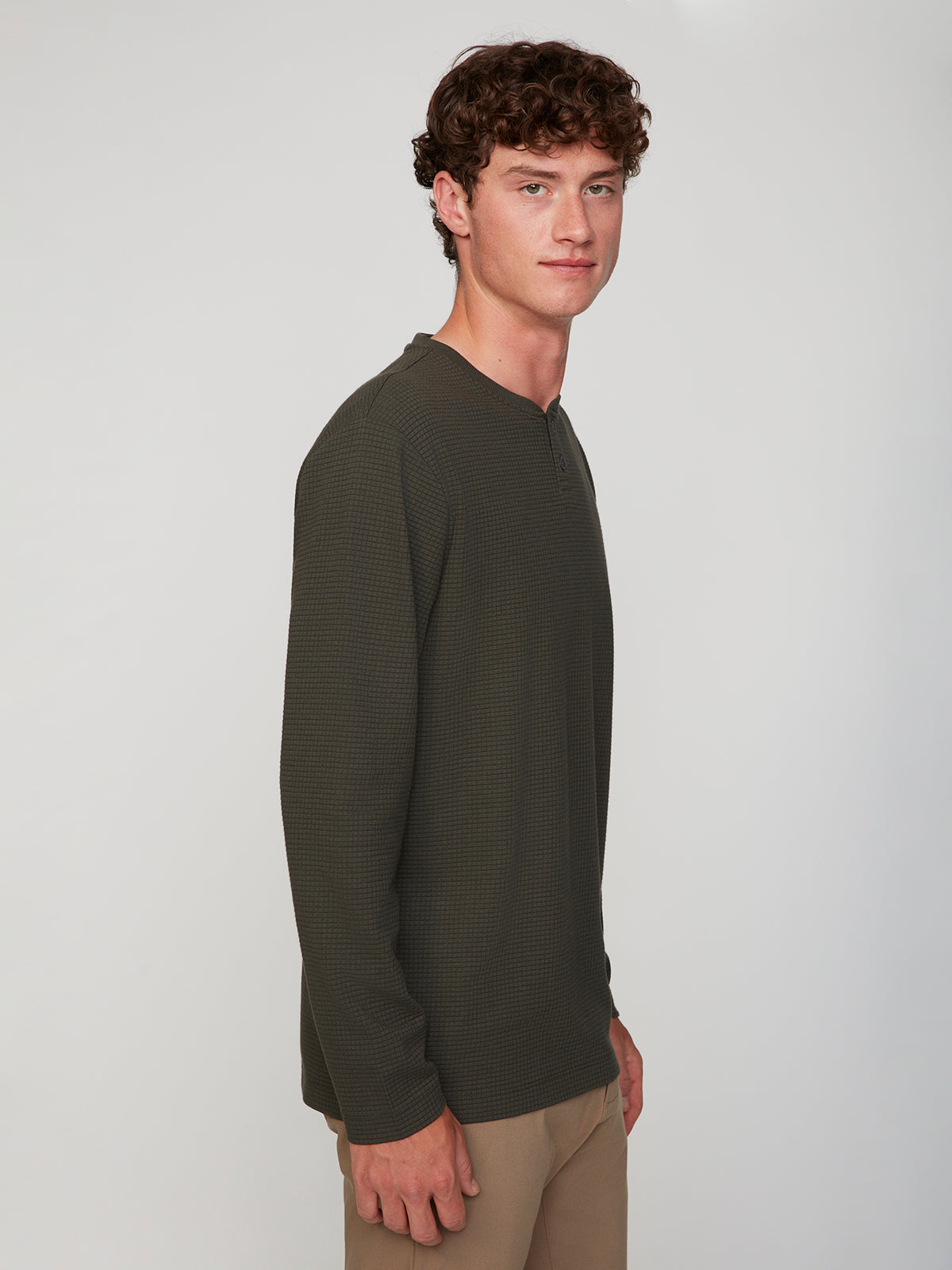 A young man with curly brown hair, wearing a dark green PROJEK RAW MENS LONG SLEEVE HENLEY GRID JERSEY and light brown pants, stands slightly turned against a plain, light background.