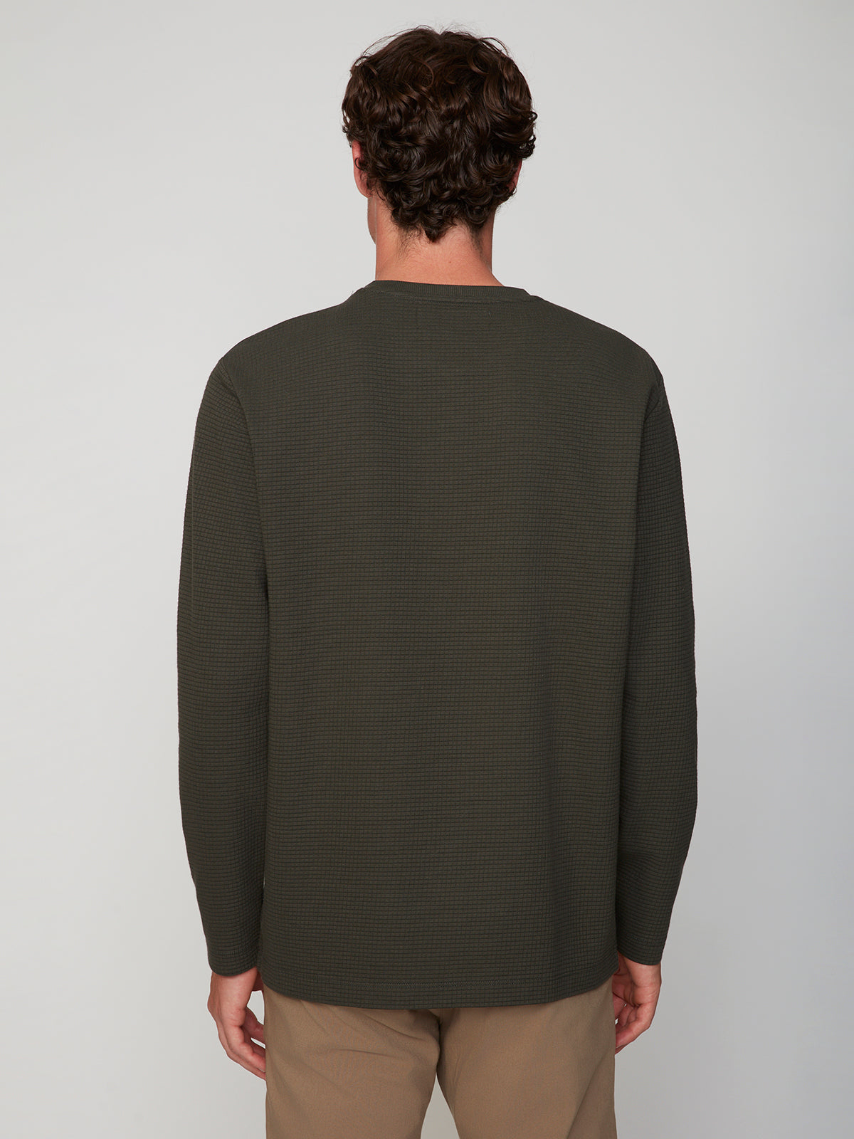 A man with short brown hair, seen from behind, wears a PROJEK RAW MENS LONG SLEEVE HENLEY GRID JERSEY in dark olive green and beige pants, standing against a plain light gray background.