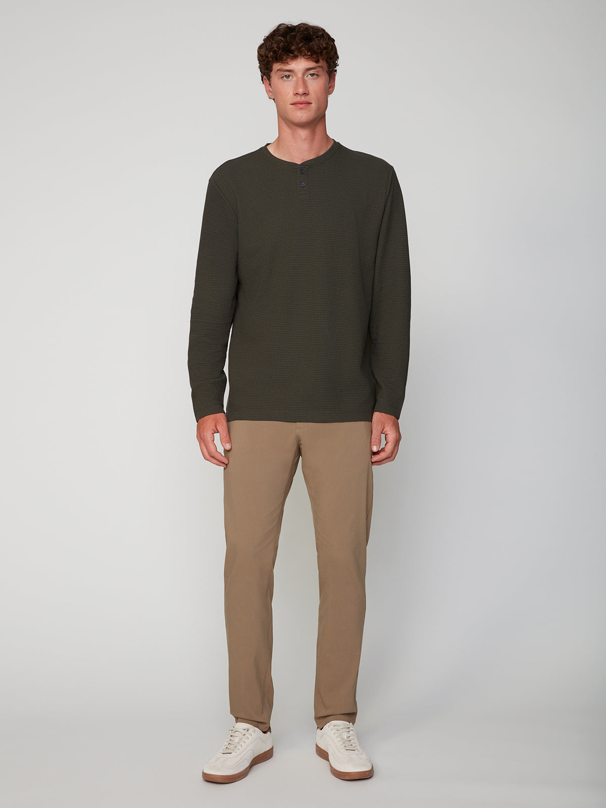 A man stands facing forward against a plain light gray background, wearing a PROJEK RAW MENS LONG SLEEVE HENLEY GRID JERSEY, light brown pants, and white sneakers.
