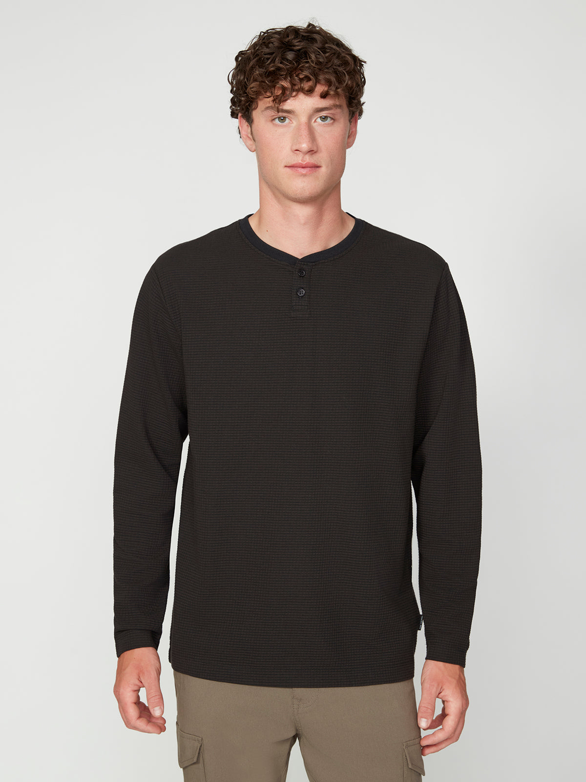A young man with curly brown hair stands facing forward, wearing a PROJEK RAW MENS LONG SLEEVE HENLEY GRID JERSEY and light brown pants against a plain white background.
