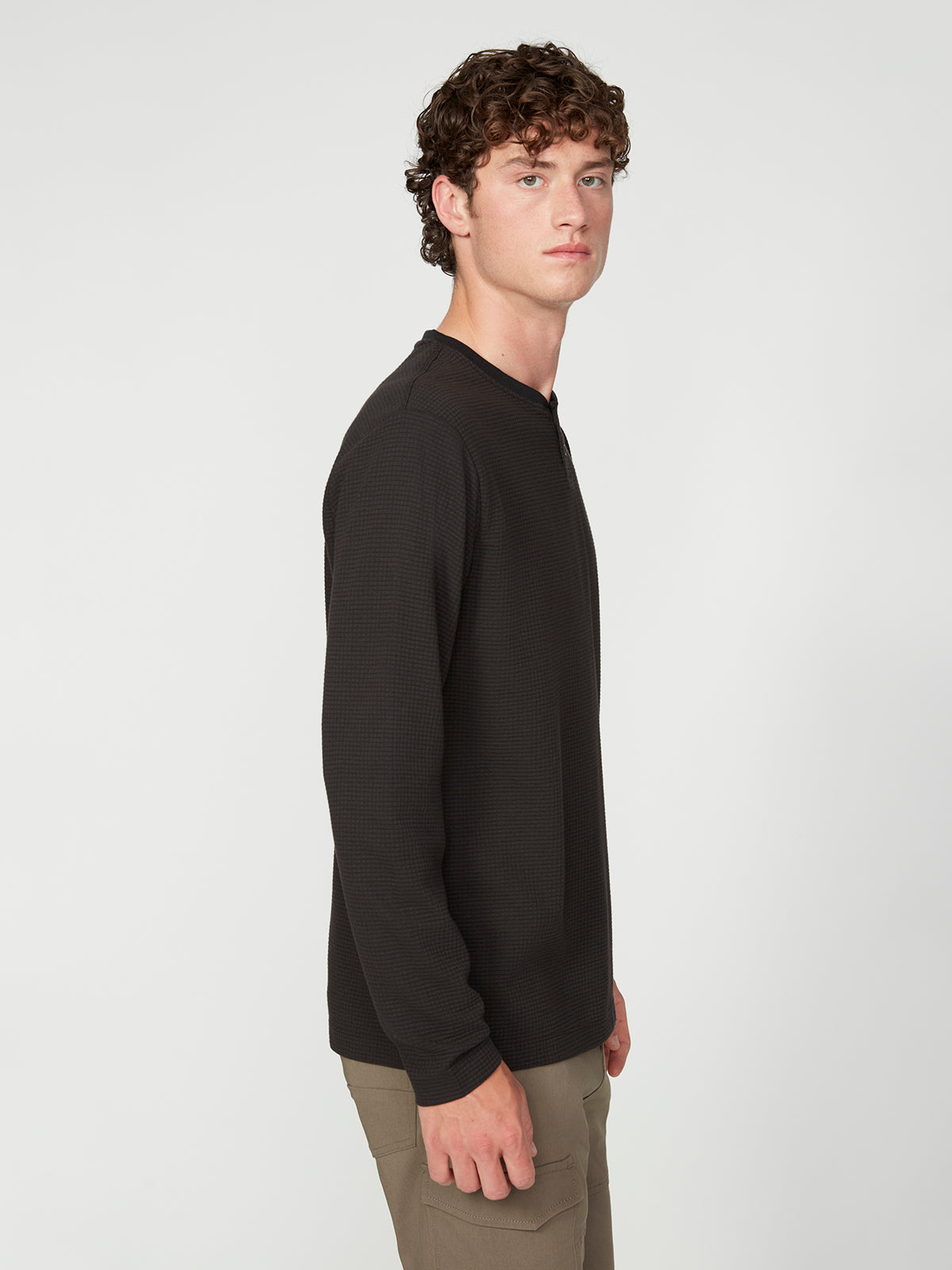 A young man with curly brown hair stands sideways, looking at the camera. He wears a PROJEK RAW MENS LONG SLEEVE HENLEY GRID JERSEY and khaki pants against a plain white background.