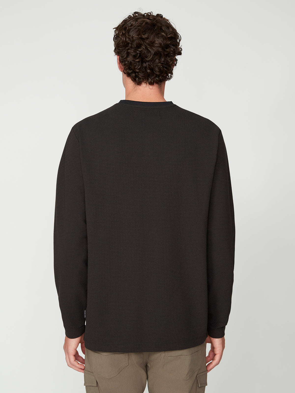 A man with curly brown hair faces away, wearing the PROJEK RAW MENS LONG SLEEVE HENLEY GRID JERSEY in black and light brown cargo pants against a plain white background.