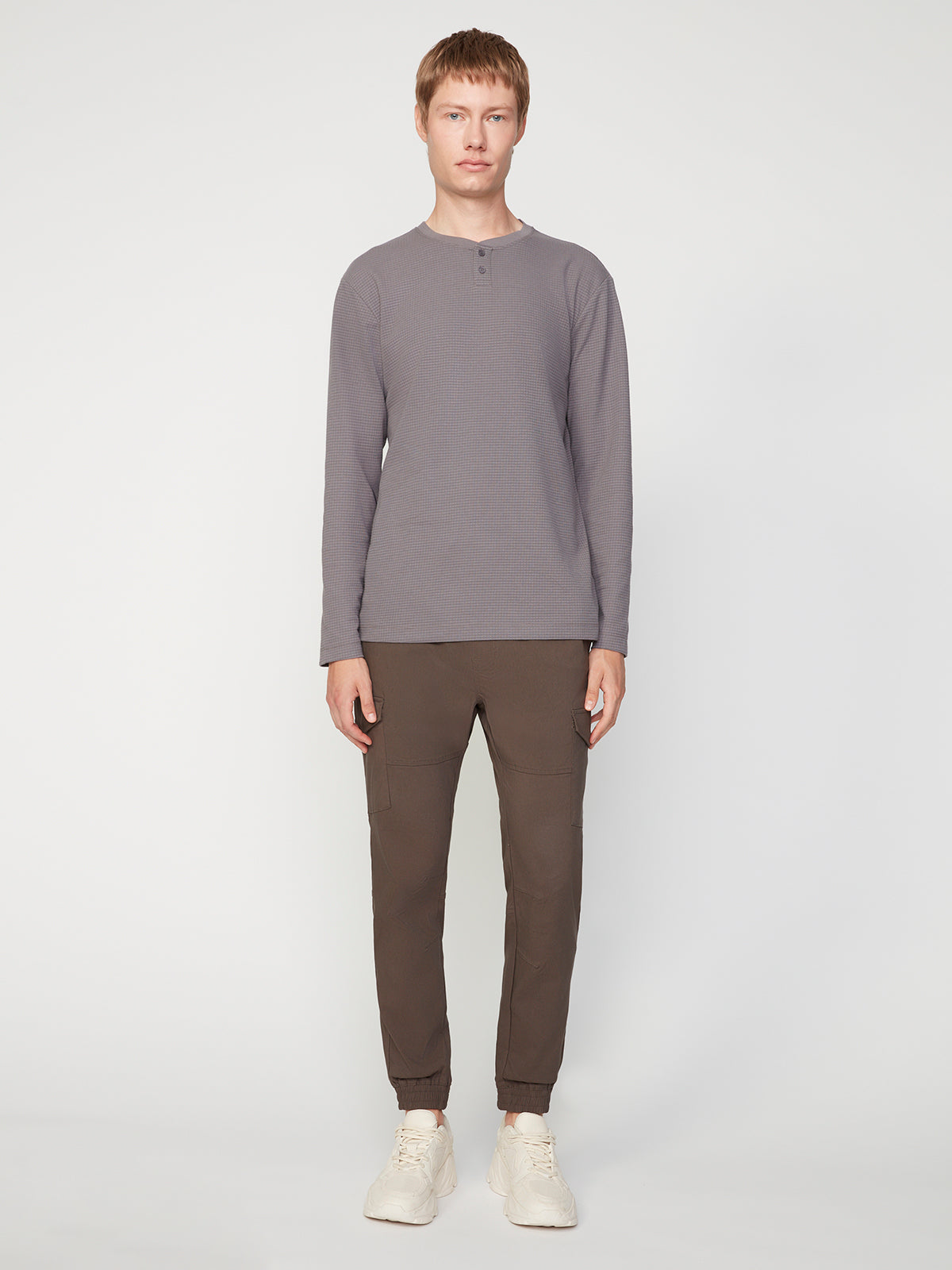 A person with short light brown hair wears a PROJEK RAW MENS LONG SLEEVE HENLEY GRID JERSEY in gray, dark brown jogger pants, and white sneakers, posing against a plain white background.