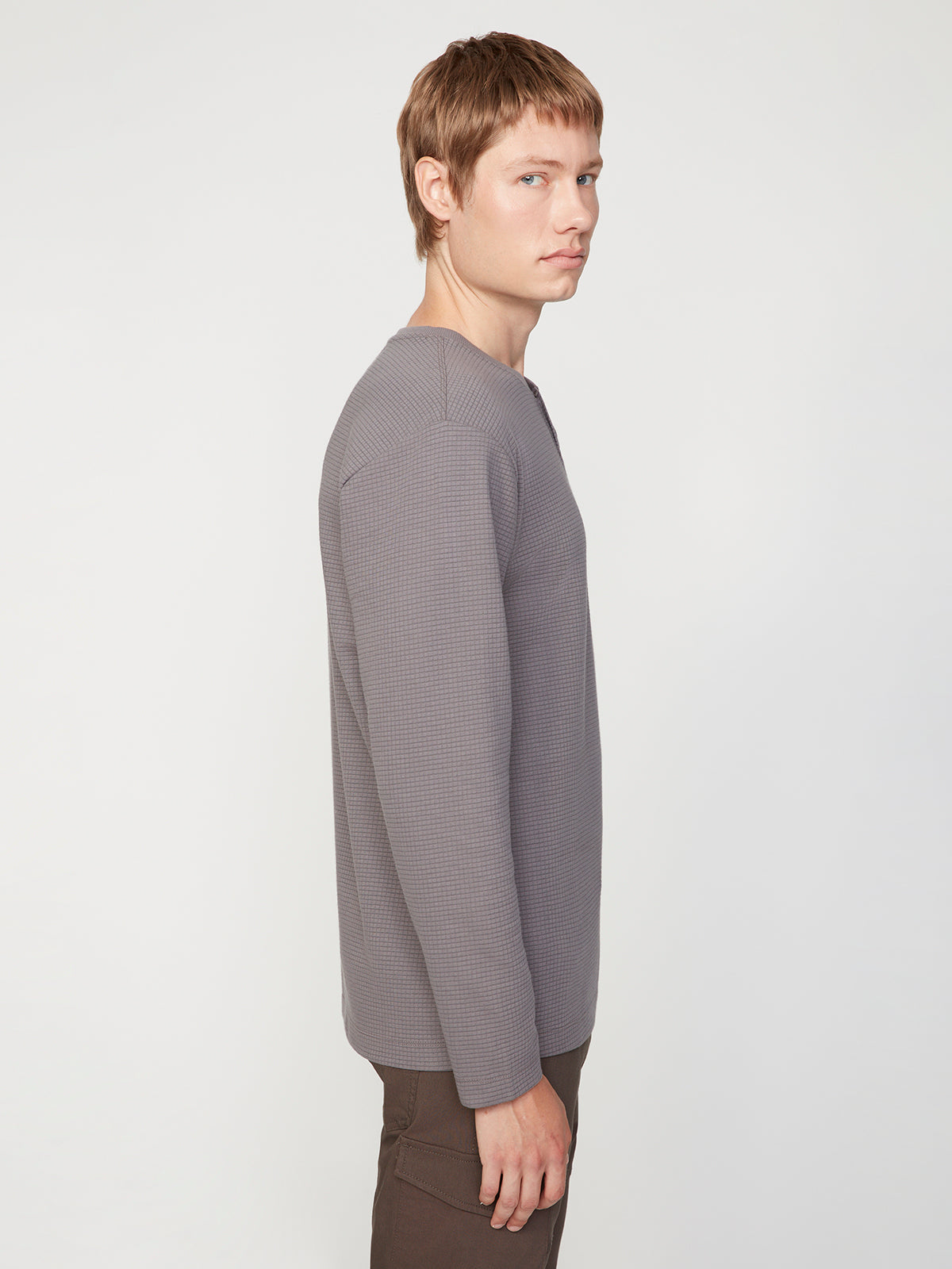 A person with short, light brown hair models the PROJEK RAW MENS LONG SLEEVE HENLEY GRID JERSEY and dark pants, standing sideways against a plain white background.