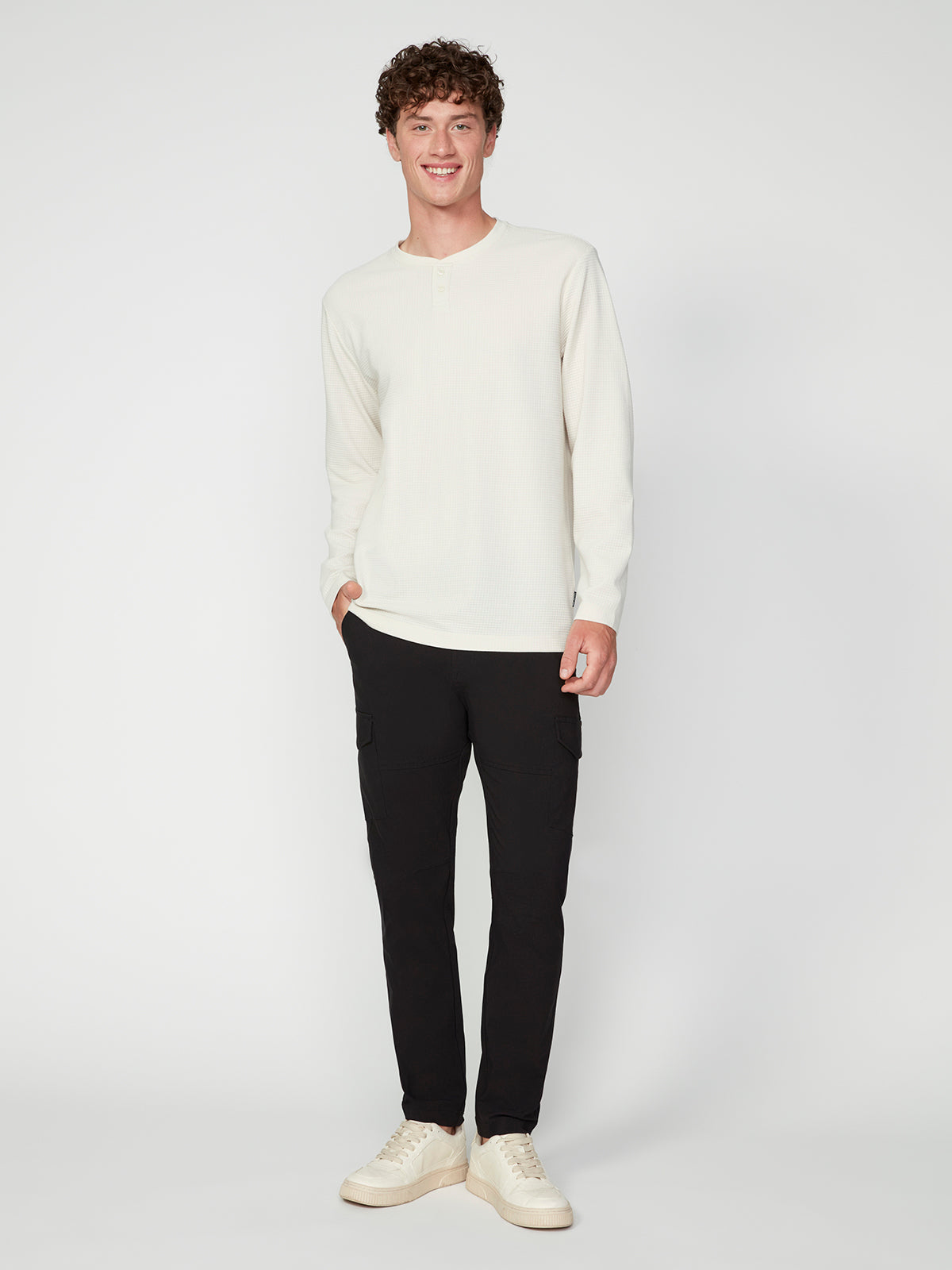 A young man smiles in a PROJEK RAW MENS LONG SLEEVE HENLEY GRID JERSEY, black cargo pants, and white sneakers, standing against a plain white background.