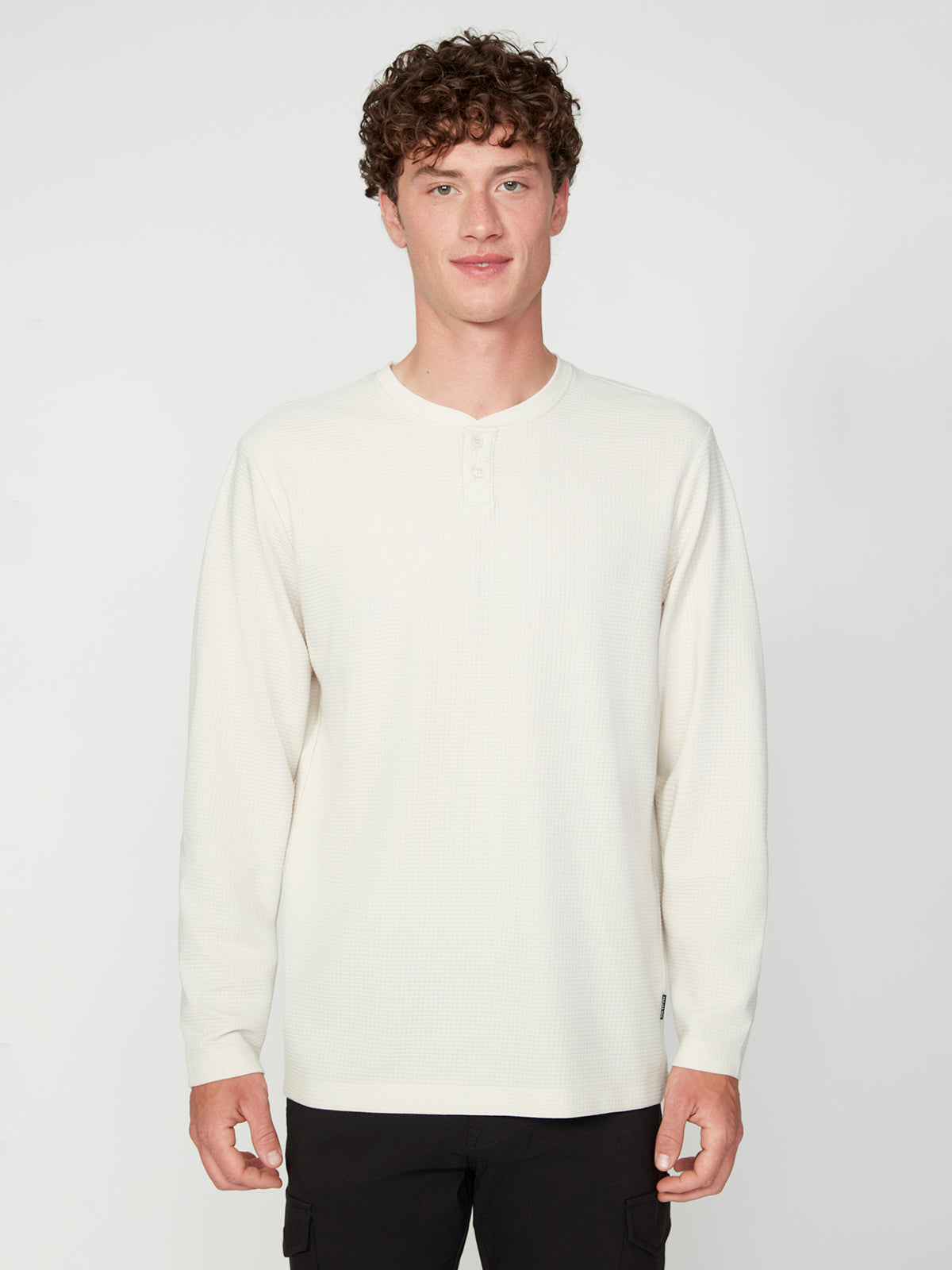 A young man with curly brown hair wears a PROJEK RAW MENS LONG SLEEVE HENLEY GRID JERSEY in off-white and black pants, standing against a plain white background and facing the camera with a neutral expression.