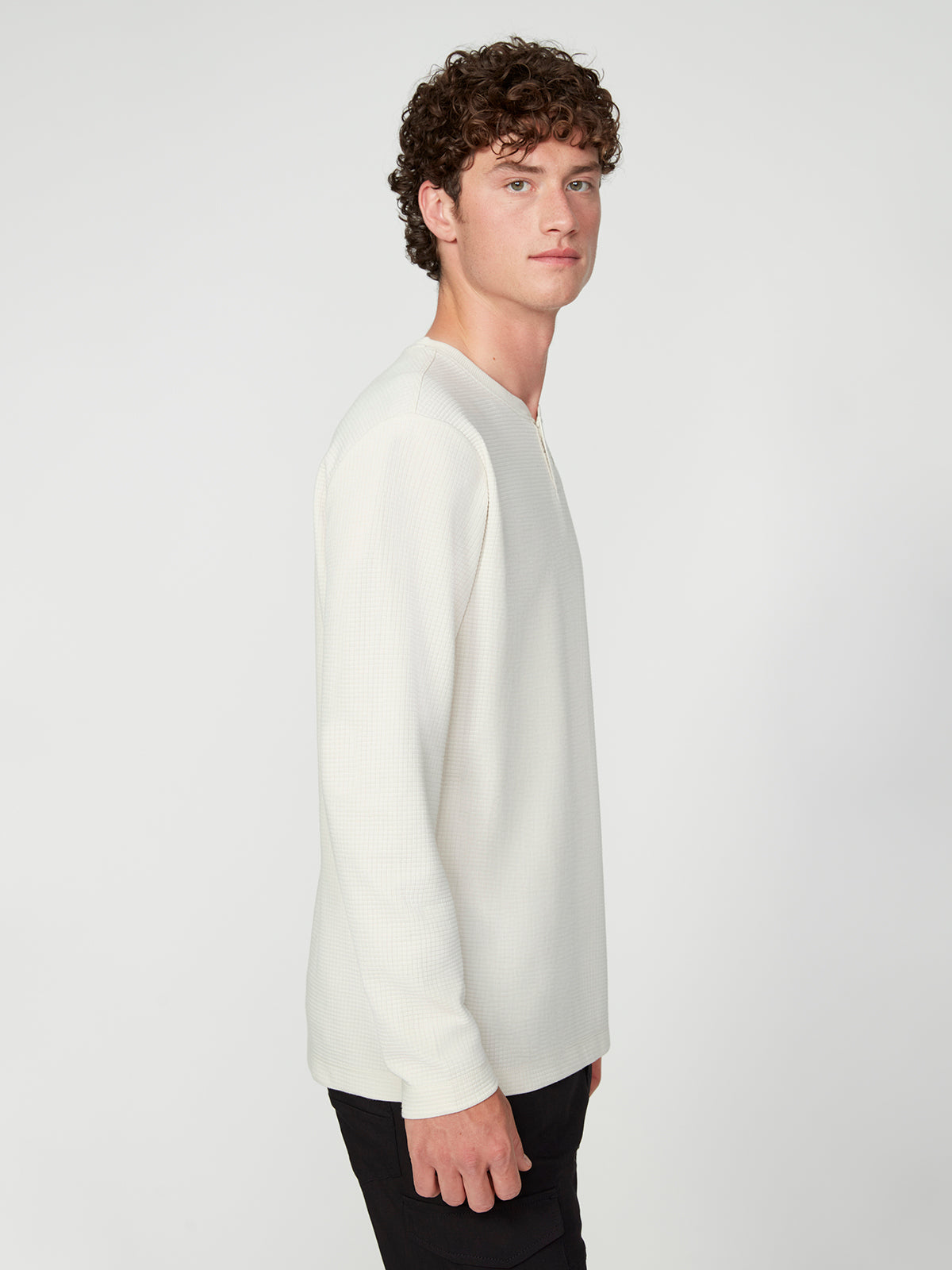 A young man with curly brown hair wears a PROJEK RAW MENS LONG SLEEVE HENLEY GRID JERSEY in off-white and black pants, standing sideways against a plain white background, looking at the camera with a neutral expression.