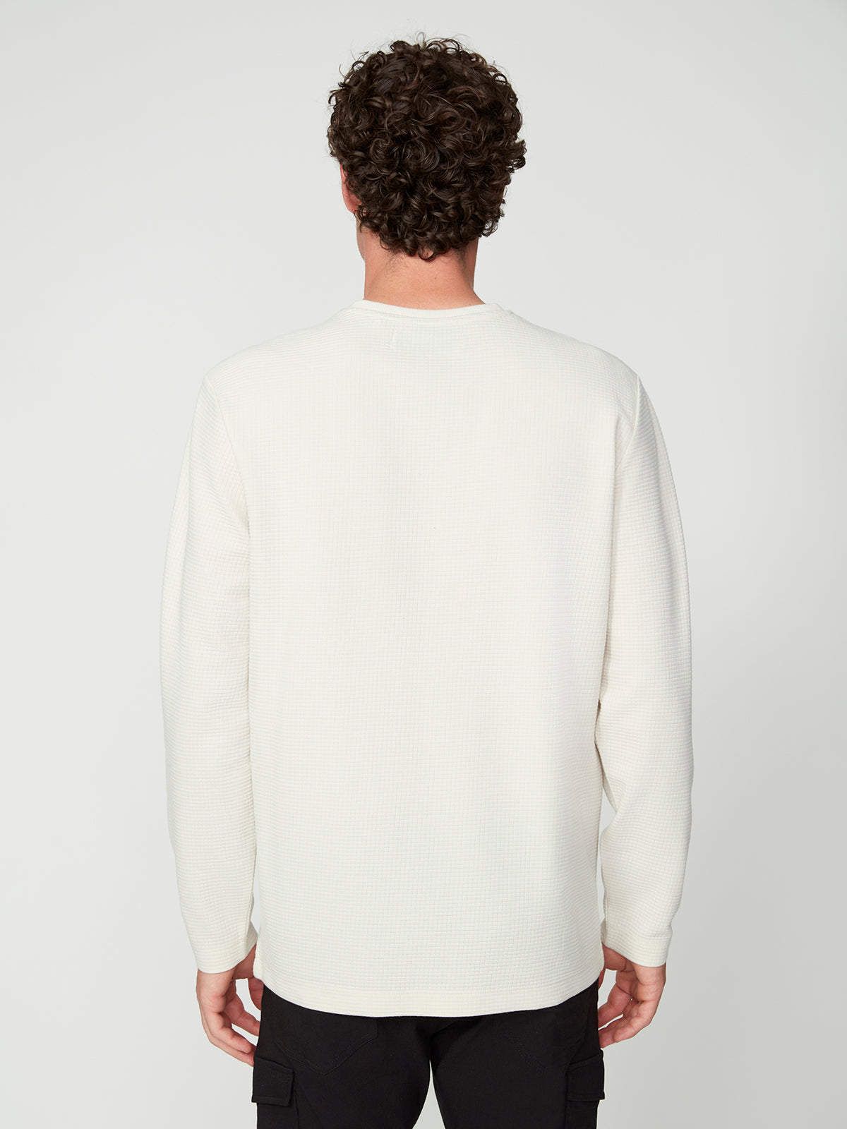 A person with curly brown hair is seen from behind, wearing the PROJEK RAW MENS LONG SLEEVE HENLEY GRID JERSEY and black pants, standing against a plain light gray background.