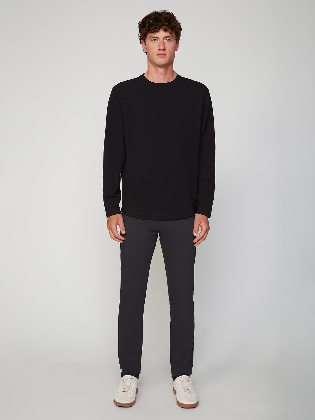 A young man with curly hair faces forward, wearing a PROJEK RAW MENS LONG SLEEVE CREW NECK, dark gray pants, and white sneakers against a plain light gray background.