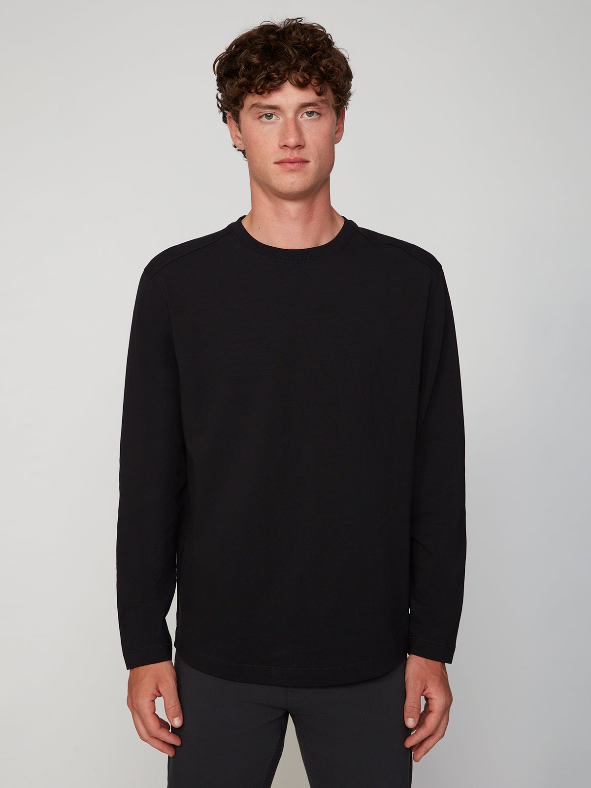 A young man with curly brown hair stands facing forward in a PROJEK RAW MENS LONG SLEEVE CREW NECK shirt and dark pants, set against a light gray background.